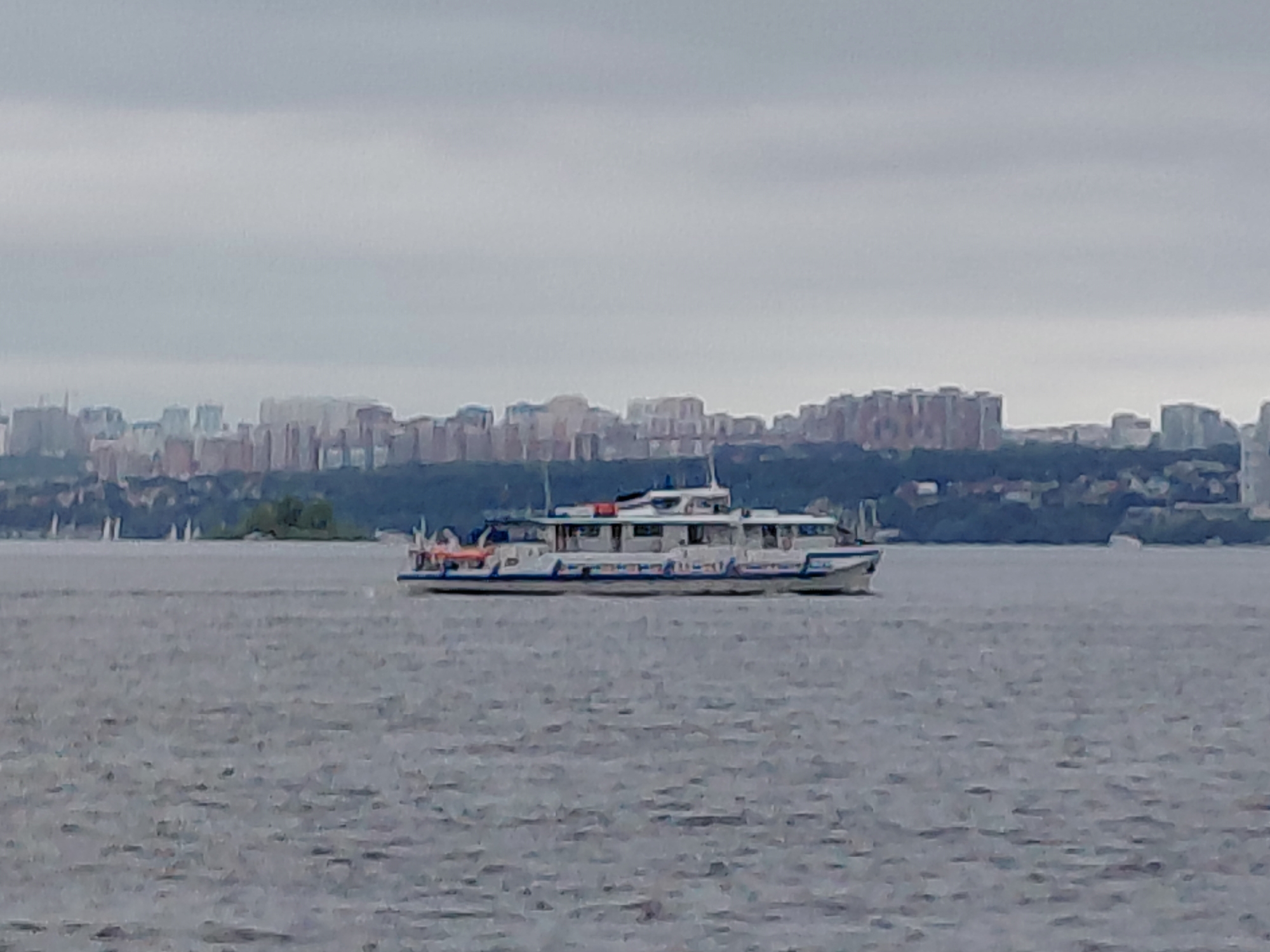 A medium shot captures a cloudy day scene over a body of water, likely a river, with a city skyline in the background. In the foreground, a white passenger boat with blue accents sails from left to right across the water. The boat has multiple windows along its length and a closed cabin area with a white roof. The water appears choppy. In the middle ground, a small, tree-covered island is visible on the left. The background features a dense cityscape with numerous buildings, ranging in height, and some greenery. The sky is overcast with gray clouds. This scene is situated in Rozhdestveno, Russia.