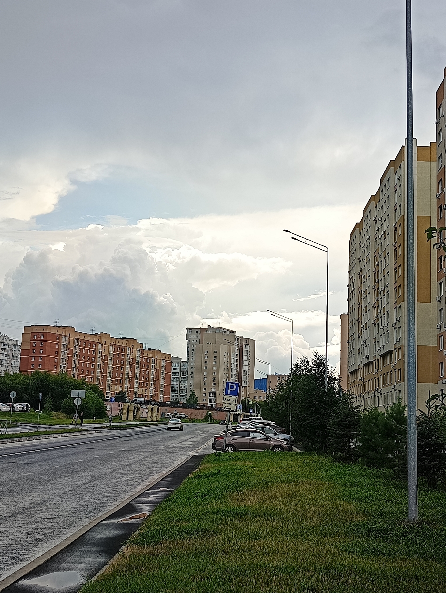 The image depicts a street scene in Kemerovo, Russia, characterized by a cloudy sky and overcast conditions. In the foreground, a paved road with wet surfaces and puddles suggests recent rain, bordered by a strip of green grass. Beyond the grass, several cars are parked on the side of the road. The middle ground features a series of multi-story residential buildings with brick and light-colored facades, typical of urban architecture. Trees are interspersed between the buildings and along the roadside. In the background, more buildings and a large, dense cloud formation dominate the sky, adding to the somber atmosphere of the scene. The image appears to be taken during daylight hours, but the cloud cover significantly reduces the natural light.