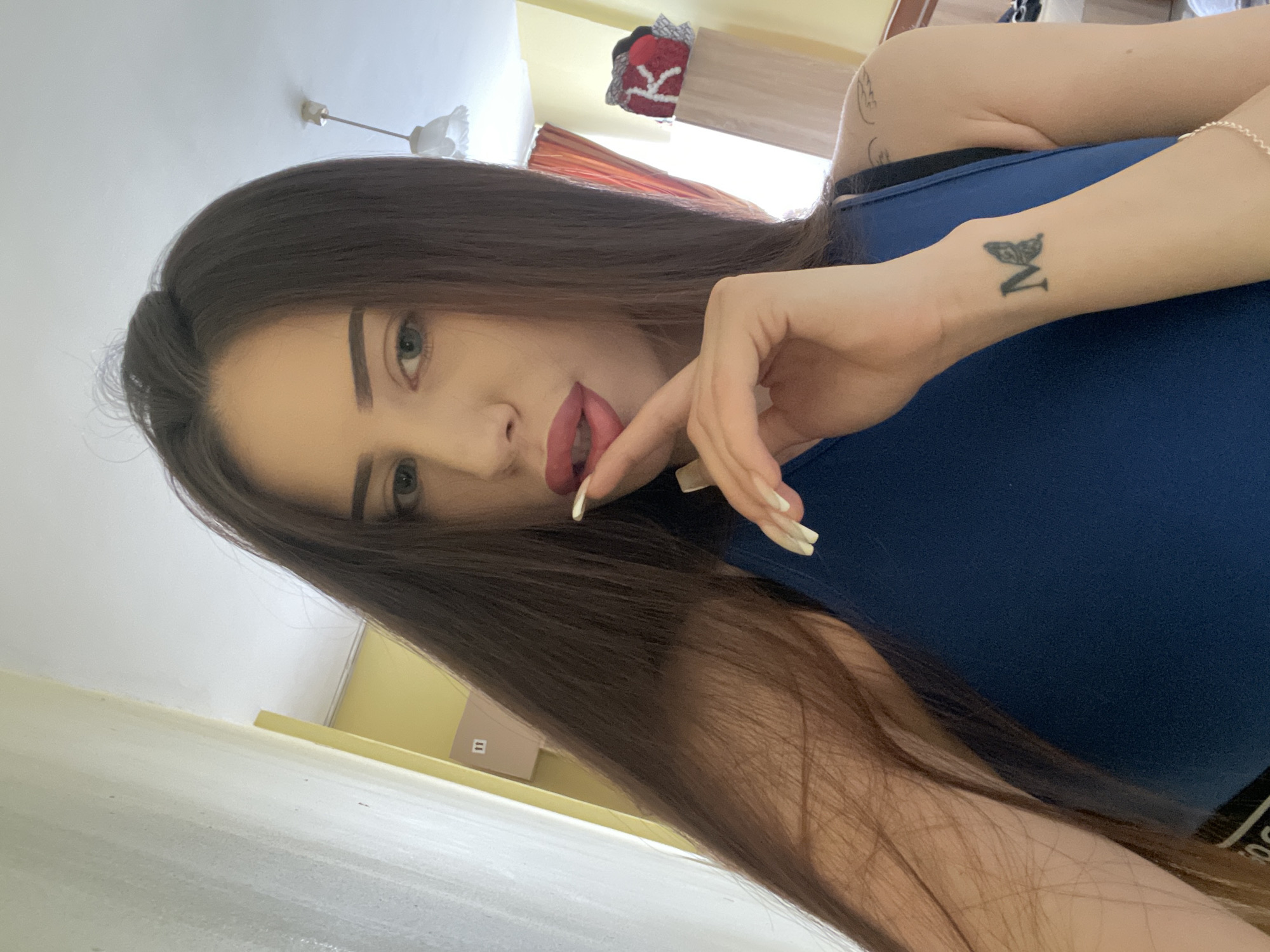A young woman with long, dark brown hair is shown in a close-up selfie. She is wearing a dark blue tank top. Her left hand is raised to her face, with her index finger touching her lips, which are painted red. She has long, pointed fingernails. A small tattoo of a butterfly with the letter "M" inside it is visible on her left wrist. In the background, a light-colored wall and ceiling are visible, along with a part of a wooden shelf with some folded items on it, including a red item with a white "K" on it. A ceiling light fixture is also visible. The scene appears to be indoors.