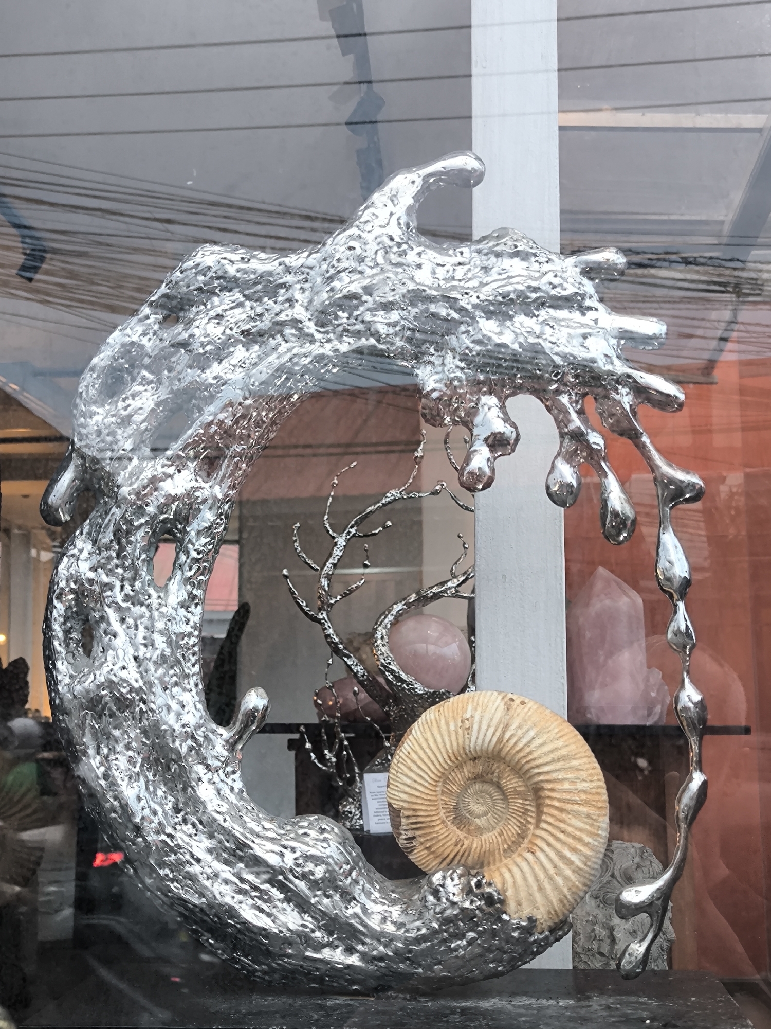 This media file captures a sculptural display seen through a window, likely in a commercial setting in North Kuta, Indonesia. The central and most prominent subject is a large, abstract sculpture crafted from shiny, silver-colored metal. It appears to depict molten liquid or a stylized wave, forming a dynamic, open crescent shape with various dripping elements and textured surfaces.
Within this display, and integrated with the silver sculpture, is a large, cream-to-brown colored ammonite fossil, showcasing its distinct spiral pattern and ribbed shell. Behind these main elements, several other objects are visible: a delicate, branched metallic piece, possibly resembling coral or a stylized tree branch, and multiple polished pink or rose quartz crystals, including columnar forms and a sphere.
The window glass reflects the external environment, providing context. Clearly visible are numerous overhead wires and cables stretching across the upper portion of the frame, alongside reflections of light-colored building facades. A strong orange-red hue dominates the background on the right side due to a reflection of an external wall or structure. Dark, indistinct shapes, possibly pedestrians or other objects, are also faintly reflected.
The arrangement suggests an artistic or retail display, possibly for a gallery or a shop specializing in unique artifacts, jewelry, or spiritual items. A small card or label is partially visible behind the ammonite, with fragments of text like "SILVER" and "SCULPTURES" discernible, indicating the material and type of some of the displayed items. The lighting suggests it is daytime. No people are directly visible within the frame, and the scene is static, showcasing the artwork.