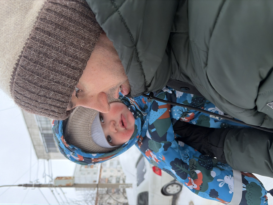 A close-up depicts a father and child in winter clothing. The father wears a tan and brown beanie and a dark green puffer jacket. The baby wears a light-colored beanie and a blue snowsuit with a woodland creature print. The child's face is visible, with rosy cheeks. A street scene with buildings and a white car is visible in the blurred background. The photo is oriented sideways.