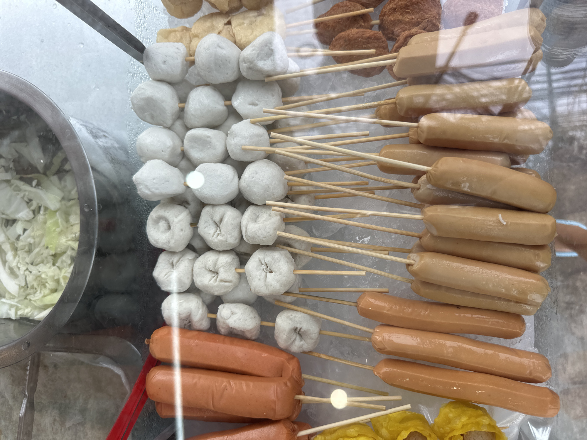 The image displays a close-up, overhead view of a food stall in Phuket City Municipality, Thailand. The stall appears to be a street food vendor, with various items displayed on skewers and in a metal container.
The primary subjects are food items arranged on wooden skewers, including white fish balls, pale brown sausages, and some fried brown items, possibly patties or more fish balls, at the top. In a circular metal container to the left, there is a pile of shredded white cabbage, likely for serving with the food. The food is laid out on a bed of ice, suggesting it is being kept cold.
The scene is brightly lit, likely indicating it is daytime, though no specific time can be determined. There are reflections on the glass or plastic covering the food, indicating the vendor is enclosed. The overall impression is of a vibrant and busy food stall, typical of street food scenes in Thailand. No people are visible in the frame. There is no visible text in the image. The activity taking place is the preparation and display of food for sale.