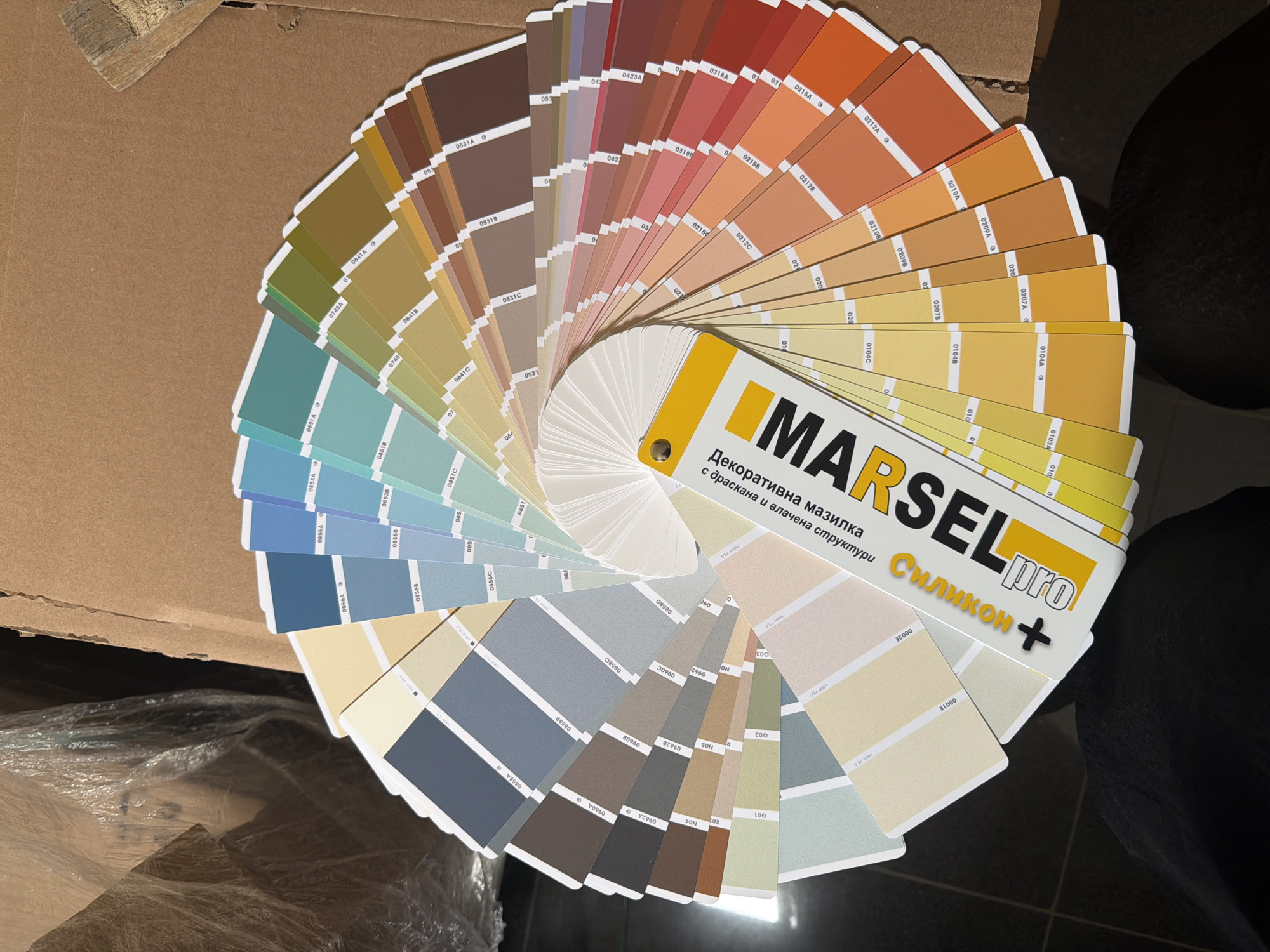 The image shows a color sample fan deck spread out on a cardboard surface. The brand "MARSEL pro" is prominently displayed on the cover of the deck, along with the tagline "Декоративна мазилка с драскана и влачена структури Силикон +". The fan deck displays a variety of colors, ranging from browns and reds at the top, transitioning through yellows and greens, and ending with blues and grays at the bottom. Color codes are printed on each sample. Part of a paintbrush is visible in the upper left corner of the image. The scene seems to be indoors, as suggested by the lighting and the presence of packaging materials in the background. The location is not apparent from the visual information in the image, so nothing about Dolno Ablanovo, Bulgaria, can be determined.
