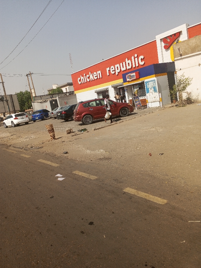 The image depicts an exterior scene in Gawuna, Nigeria, featuring a "Chicken Republic" fast-food restaurant. In the foreground, a dark asphalt road with faded yellow dashed lane markings contains scattered white paper and dark stains. To the right of the road, a dirt and gravel shoulder transitions to a parking area. A short, thick wooden post or stump stands on this shoulder.
In the midground, a dark red crossover SUV is parked facing left on the dirt lot. A dark-skinned individual wearing a white shirt and dark pants walks left, carrying a white bucket in their left hand, positioned next to the red SUV. Beyond this vehicle, several other cars are parked in the unpaved lot, including a white sedan, a blue sedan, and a dark-colored sedan.
The background is dominated by the single-story "Chicken Republic" building, which features a prominent red fascia with "chicken republic" in large white lettering. A yellow and white "Y" logo is visible on the upper right section of the building. The entrance area is recessed, with blue and yellow architectural accents. Several individuals are visible near the glass entrance doors, and a promotional sign for "SHAWARMA & SHAWARMA SNACKS" is displayed to the right of the entrance. A small green shrub grows near the building's right side. Utility poles with overhead lines are present on the left side of the scene, and a communication tower is visible in the distant background under a clear, light blue sky. The overall environment appears dry and dusty.