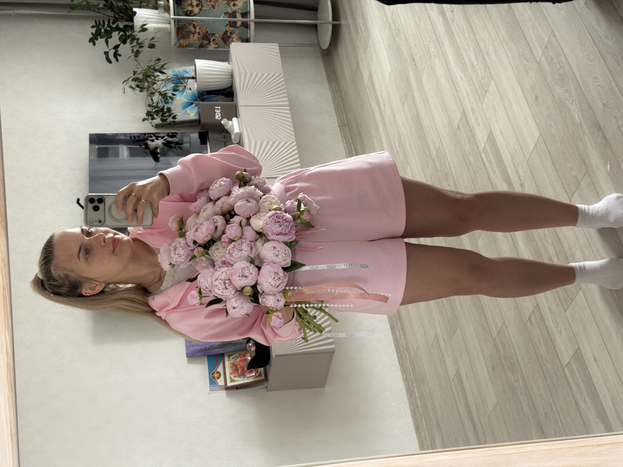 An indoor, full-body mirror selfie captures a female in Chaikovsky, Russia. The subject, a Caucasian female estimated to be in her late 20s to early 30s, stands centered within the mirror's reflection. She has long blonde hair tied in a high ponytail and wears a pink hooded sweatshirt, matching pink shorts, and white ankle socks. A visible ear piercing is present on her left ear's cartilage. She holds a large bouquet of numerous light pink peonies with both hands. Multiple ribbons, including a pink satin ribbon, a white ribbon with "ILSI" text, and a string of faux pearls, hang from the peony stems. Concurrently, she holds a silver-colored smartphone, consistent with an iPhone Pro model due to its visible camera array and a circular MagSafe accessory, in her left hand, positioned to capture the reflection. Her gaze is directed slightly to her right, towards the reflected phone screen, with a neutral to mildly pleasant facial expression.
The background, visible in the mirror, consists of a light off-white wall. A light-colored console table or cabinet featuring two doors with a radial, sunburst-like etched pattern extends horizontally across the mid-ground. From left to right on the console, visible objects include partially obscured framed items (one appearing devotional or artistic), a small black object, a small white ceramic figurine, a dark grey rectangular digital clock displaying the reflected numerals "20:E1" (indicating 13:02), a small green potted plant in a white ribbed pot, a larger green potted plant also in a white ribbed pot, and a multi-tiered display unit to the far right holding multiple framed images of domestic cats and kittens. The floor is covered with light-toned wooden or laminate planks arranged in a staggered pattern.