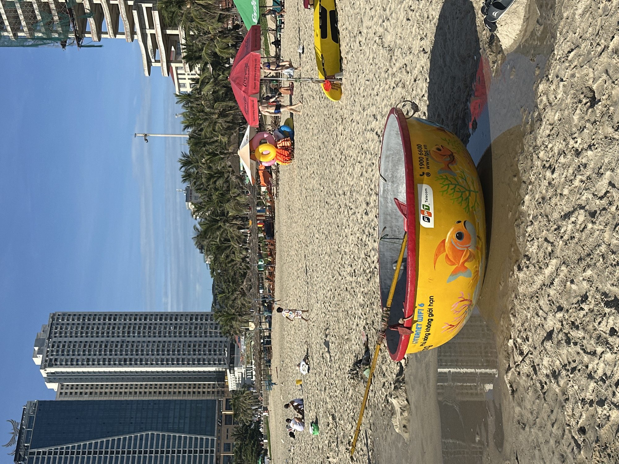 The image captures a scene at My Khe Beach in Da Nang, Vietnam. The foreground features a yellow, circular basket boat with red trim, partially submerged in shallow water on the sandy beach. The boat is adorned with a painted image of an orange goldfish and green coral, along with the FPT Telecom logo and contact information. Two yellow oars are positioned across the boat.
In the midground, the sandy beach is populated with several people enjoying the day. Some individuals are seated, while others are standing or walking. Further back, a child is running across the sand. Beach umbrellas, including a red one with a Coca-Cola logo and a green one, provide shade for beachgoers. Beach chairs and what appear to be inflatable water toys are visible near the umbrellas.
The background is dominated by the urban landscape of Da Nang. Several tall, modern buildings, including skyscrapers, are visible along the coastline. One building appears to be under construction, with scaffolding and netting. Palm trees line the beach, adding to the tropical atmosphere. The sky is clear and blue, indicating fair weather. The overall impression is a vibrant beach scene with a blend of natural beauty and urban development.