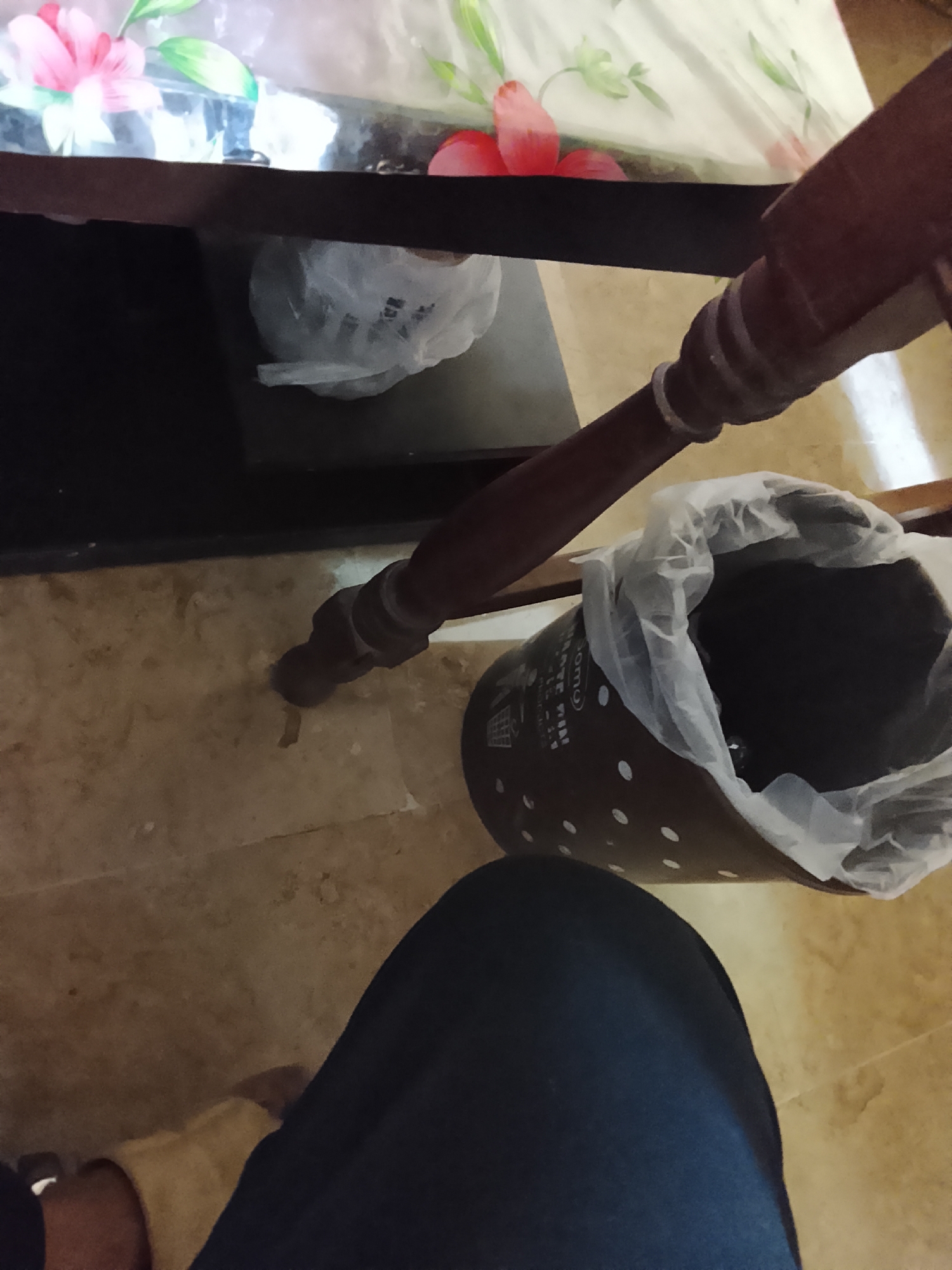 The scene depicts a partially visible person wearing dark blue pants, seated near a brown wooden table with a floral patterned top. A trash can with white polka dots and a plastic bag liner sits on a light brown tiled floor near the table leg. Another plastic bag is visible under the table. The specific location in Multan, Pakistan cannot be determined from the visual data.