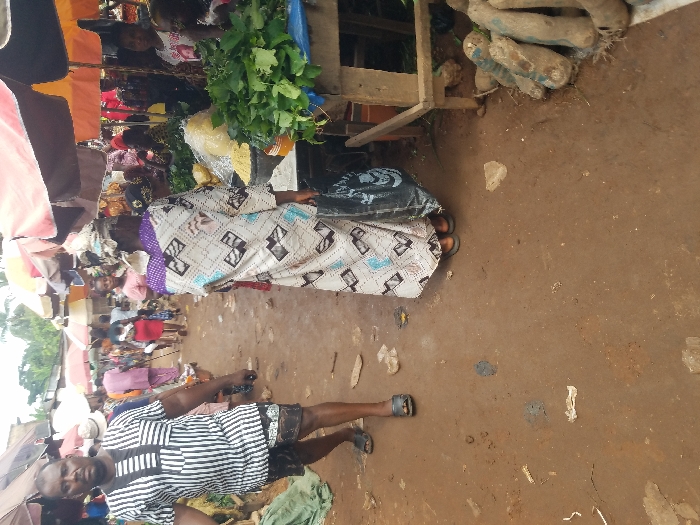 This image captures a bustling outdoor market scene in Nnobi, Nigeria. The foreground features a reddish-brown dirt path scattered with minor debris.
On the left, a dark-skinned male subject walks towards the viewer, slightly off-center. He has short dark hair and a mustache, wearing a short-sleeved, collared shirt with alternating black and white vertical stripes and a geometric pattern on the chest. He wears patterned dark shorts and dark open-toed sandals.
In the center, a female subject is viewed from behind, walking away from the camera. She wears a patterned headwrap with purple accents and a loose-fitting, long-sleeved dress or gown in a light color with a repeating geometric pattern in black, white, and light blue. She carries a dark shopping bag in her left hand and wears dark open-toed sandals.
To the right, a rustic wooden market stall displays produce. A large pile of fresh green leafy vegetables rests on a blue cloth. To their left, a pile of yellow granular substance, possibly garri, is visible in a clear plastic bag. On the ground next to the stall, several elongated, light brown tubers, likely yams, are arranged, partially covered by a white cloth.
The midground and background show numerous other market stalls with colorful large umbrellas providing shade, predominantly in red, orange, brown, and beige. Many people are visible moving through the market, indicating active commerce. Green foliage and tree tops are discernible above the market structures in the distant background. The overall lighting is soft and even, consistent with an overcast day.
