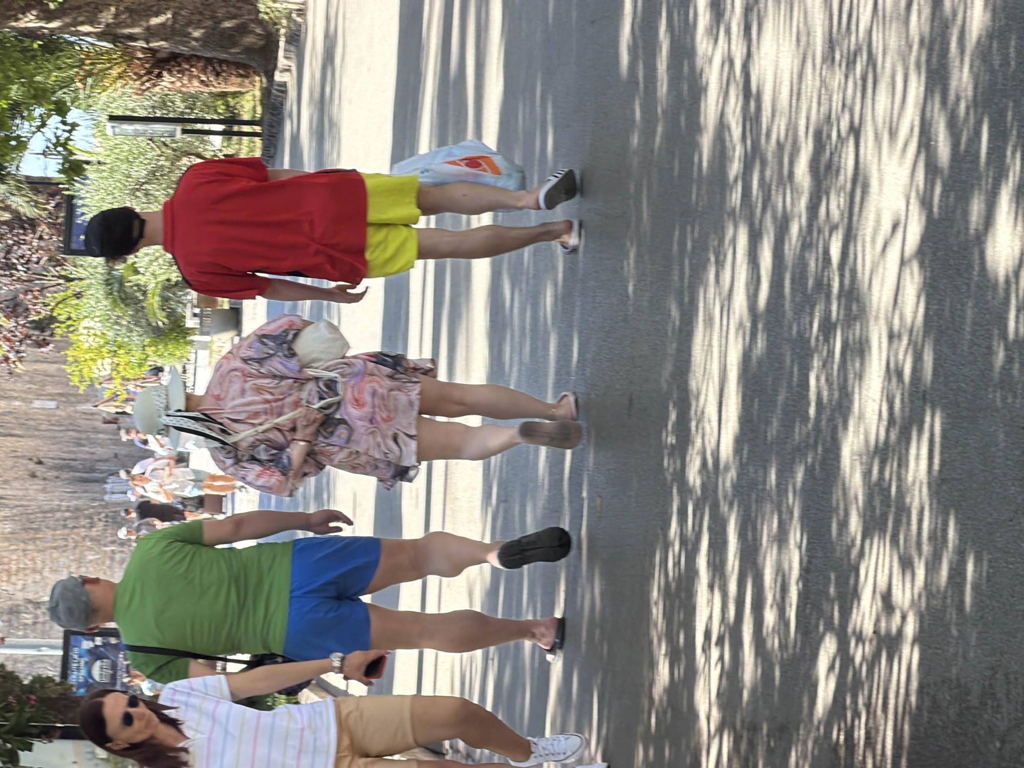 This media file captures a sun-dappled scene with several people walking outdoors, likely on a paved walkway. The orientation of the image is rotated 90 degrees counterclockwise.
The foreground shows the legs of three individuals. The first person, from the top of the frame, wears a red t-shirt and bright yellow shorts, with a black cap. They have on black and white striped athletic shoes. A white plastic bag with an orange and red logo is visible near their feet.
Below them, a person in a patterned dress with pink, purple, and white hues walks, wearing light-colored sandals. They are carrying a light-colored bag over their shoulder.
The lowest visible person in the foreground is wearing a green t-shirt and blue shorts. They have on sunglasses and a grey cap, and appear to be holding a red smartphone. A watch is visible on their left wrist. A woman in a striped shirt and tan shorts is walking in front of them, also wearing sunglasses.
In the background, other people are seen walking, some in casual attire. The setting appears to be a public area, possibly a promenade or a street lined with trees. The sunlight filtering through the leaves creates a pattern of light and shadow on the ground, suggesting it is a bright, sunny day. The foliage indicates a warm climate. No specific emotions are discernible from the image. No legible text is visible. The context provided suggests this scene is in Budva, Montenegro.