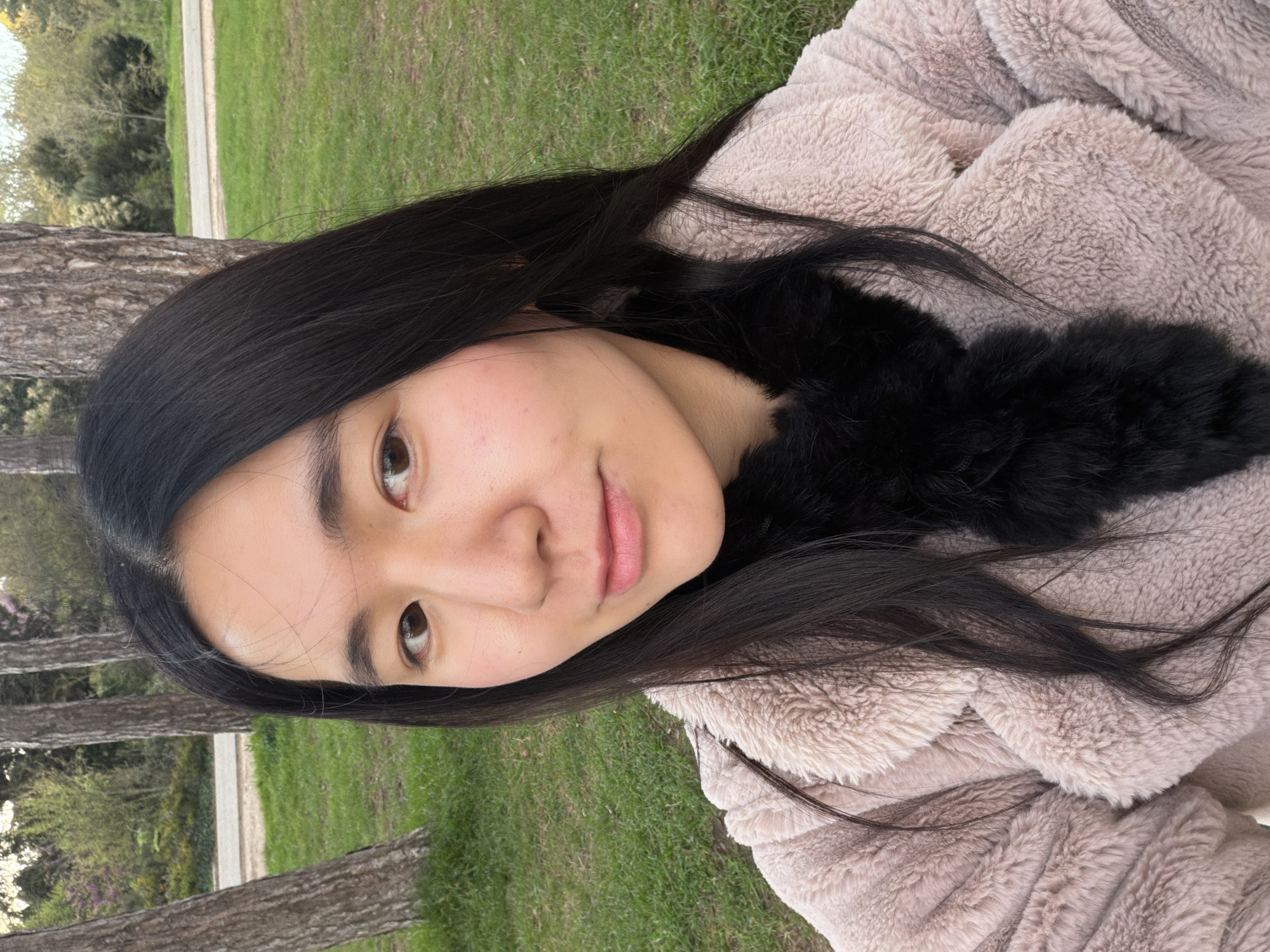 A young woman is photographed outdoors. She is wearing a beige furry coat with a black fur collar. She has long, dark hair and light brown eyes. The background consists of green grass and trees, suggesting a park-like setting. Given that the city of Paris, France, was mentioned, this might be a park located within Paris. The photograph is oriented vertically.