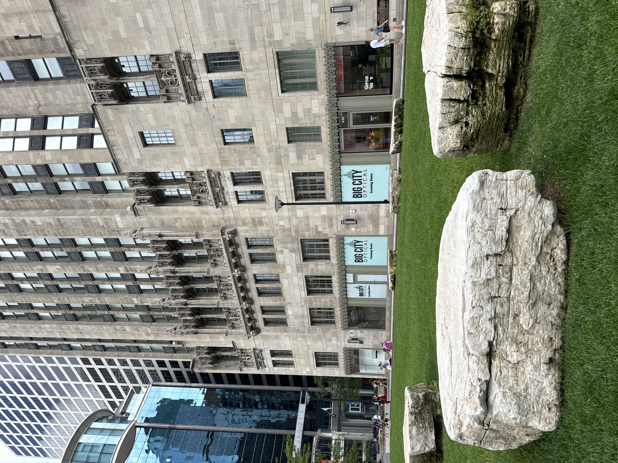 This is a vertical, eye-level, full shot of a grassy area with large rocks in the foreground, with a classic stone building and a modern glass building in the background. The grassy area is well-maintained and appears to be part of a park or urban plaza. Two large, light-colored boulders are the most prominent objects in the foreground, with a third smaller boulder visible to the left. The larger boulder on the right has a rough texture and appears weathered, with some green moss or lichen on its surface.
Behind the rocks, a tall, ornate stone building stands prominently. Its facade is characterized by a repetitive pattern of windows and intricate architectural details, suggesting it is an older, possibly historic, structure. Several signs are visible on this building, including two identical signs that read "BIG CITY OPTICAL Coming Soon".
To the left of the stone building, a modern glass skyscraper rises, its reflective surface showing the sky and surrounding buildings. The contrast between the old and new architecture is striking.
Several people are visible in the scene, mostly in the background near the buildings, suggesting a daytime setting. Some appear to be walking, while others are engaged in activities that are not clearly discernible due to the distance and image orientation. The weather appears to be overcast or the light is diffused, as there are no harsh shadows. The overall impression is an urban environment where nature, represented by the grass and rocks, has been integrated with architecture. The presence of the "Coming Soon" signs hints at a developing commercial area.
