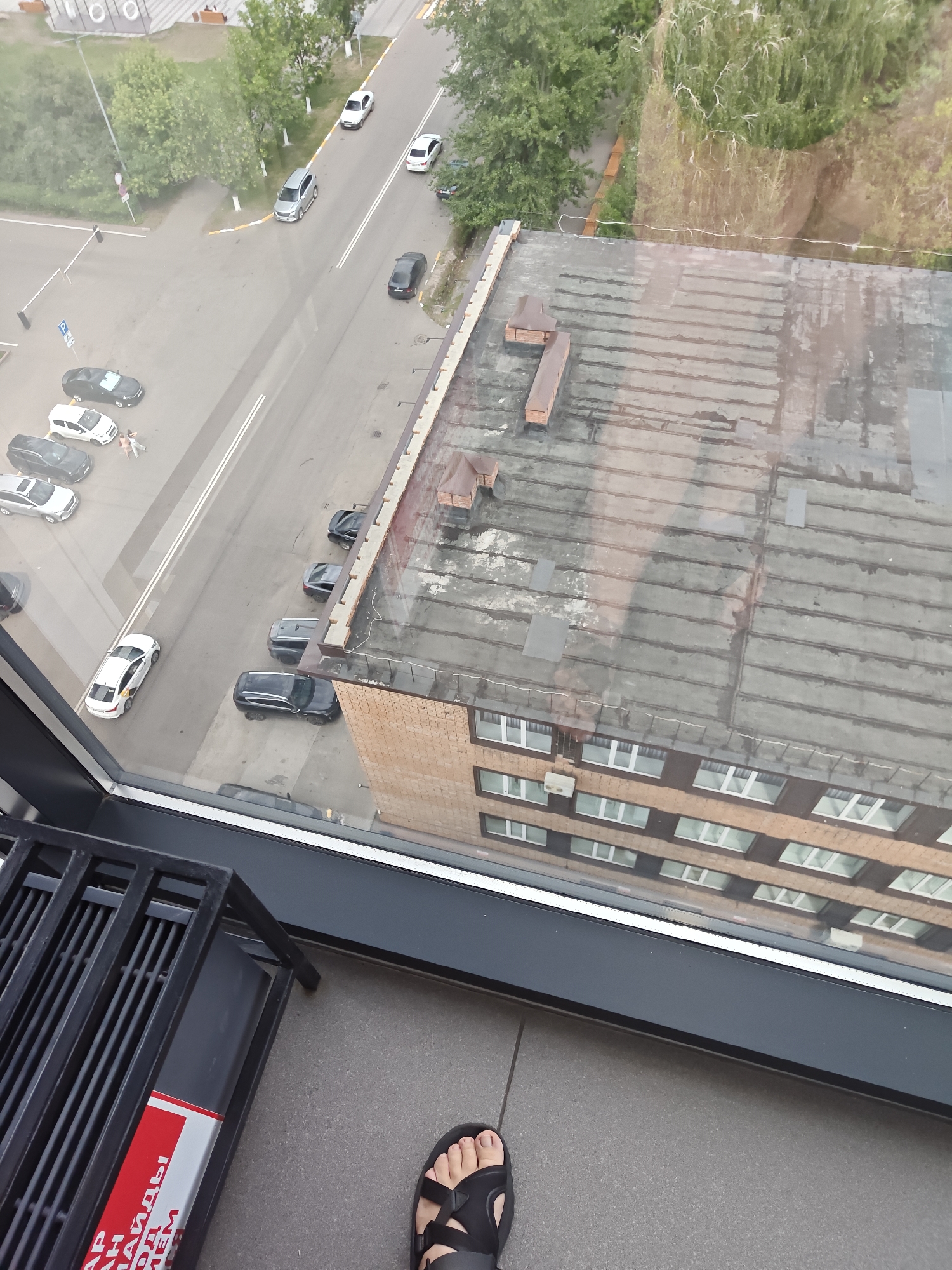 The image is a high-angle shot taken from a building, looking down at a street and an adjacent building in the city of Petropavl, Kazakhstan. In the foreground, a portion of a black metal grate and a gray tiled floor are visible. A person's left foot, wearing a black sandal, is also in the foreground.
The midground shows a street with several parked cars and moving vehicles. To the left of the street, there are parking areas and a grassy area with trees. To the right of the street, a multi-story brick building with numerous windows is prominent. The roof of this building appears to be flat and weathered, with some visible rooftop structures. Reflections of the sky and surrounding environment are visible on the windows of the building.
The background shows more trees and a glimpse of other buildings, suggesting an urban or semi-urban setting. The overall lighting suggests it might be daytime, possibly overcast or late afternoon, given the muted colors and lack of harsh shadows.