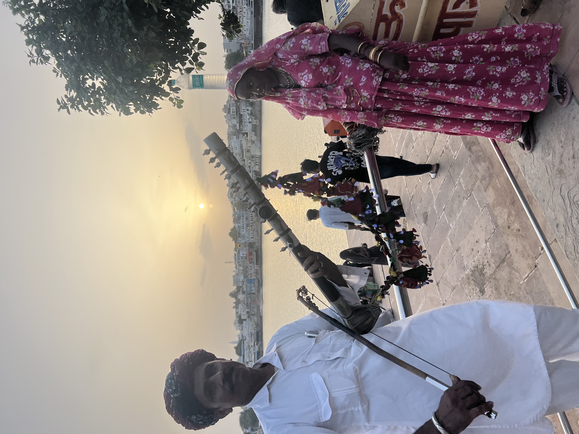 In Pushkar, India, at sunset, a man dressed in a white kurta and a colorful turban holds a bowed string instrument, possibly a Ravanahatha. He stands to the left of a woman in a bright pink floral sari and headscarf, who is adorned with bangles. They are positioned on a paved walkway overlooking a body of water, likely Pushkar Lake, with the city's buildings forming a backdrop. The sky is a pale orange and yellow, indicative of the setting sun. Several small birds are perched on the man's instrument. In the background, other people are visible, and a water bottle is placed on a ledge near a tree. The scene appears to be a public area, possibly a ghat, in Pushkar.