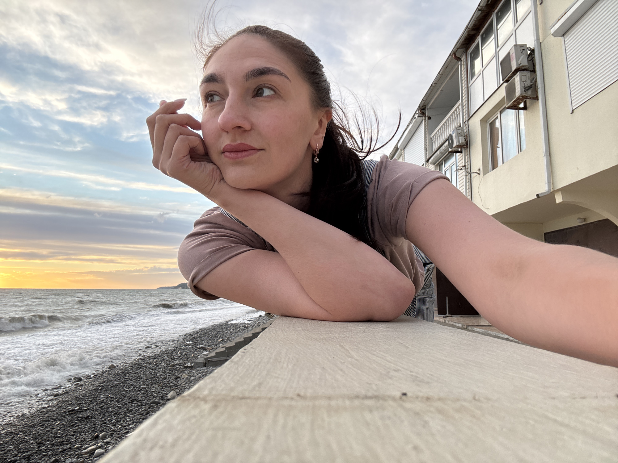 A woman poses for a selfie at a beach in Sochi, Russia. The woman is light-skinned with dark hair. She is wearing a mauve-colored t-shirt and a pair of overalls. She is resting her face on her hand, looking off into the distance. Behind her is the sea, a rocky beach, and a row of buildings. The sky is overcast with a sunset on the horizon.