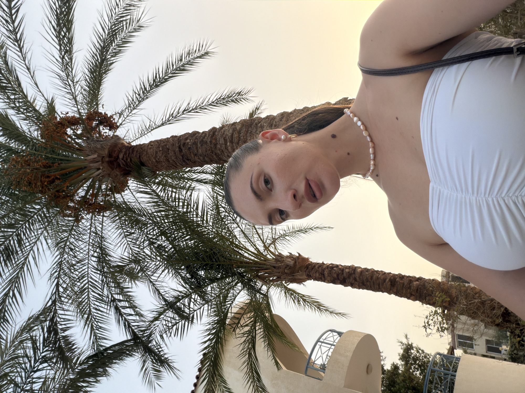 The image shows a woman outdoors, with palm trees and a building in the background. The woman is wearing a white strapless top and a pearl necklace. She has her hair pulled back and is wearing earrings. The building appears to be a hotel or resort. The scene is set during golden hour, with warm light illuminating the palm trees and the sky. This is a candid shot, likely taken on a mobile phone by the woman herself, or a companion. The image captures a moment of leisure and relaxation. The presence of palm trees and the architecture suggest a warm, possibly tropical or sub-tropical environment, such as Sharm El Sheikh, Egypt, a popular tourist destination known for its resorts and beaches.
