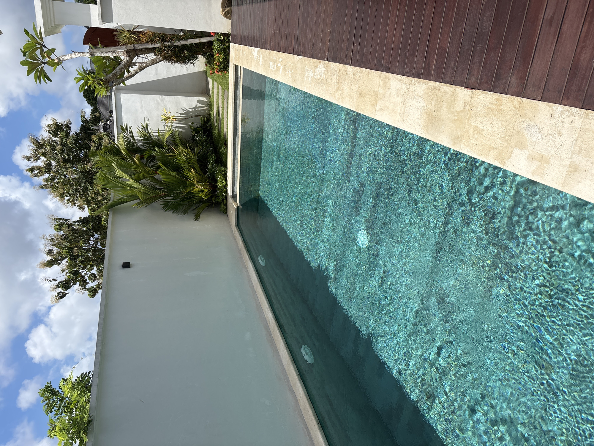 The image captures a swimming pool in Ungasan, Indonesia. The pool's water appears turquoise and ripples with sunlight. A wooden deck borders one side of the pool, separated by a light-colored stone border. On the opposite side, a white wall runs alongside the pool, partially obscured by tropical foliage, including palm trees. The wall is adorned with a small, square dark fixture. A glimpse of the sky reveals blue patches and fluffy white clouds. Other buildings and vegetation appear in the background.
