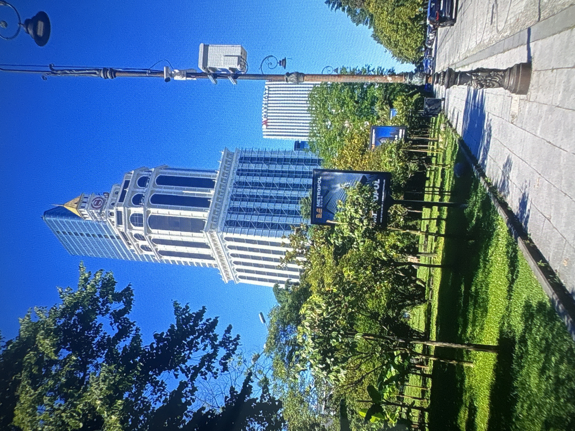 This image depicts an outdoor urban scene with a bright blue sky. In the foreground, a street lamp with decorative elements is visible, along with overhead wires. A tall, modern building with a white facade and numerous windows dominates the left side of the frame, with a gold-colored spire on its left. Adjacent to this building is another modern high-rise with a striped facade.
The scene is lined with lush green trees and a neatly mowed lawn. Several billboards are placed in the area, one of which displays the text "METROPOL CUBE". In the background, more trees and buildings can be seen. The perspective suggests a bright, sunny day, possibly midday. The image appears to be taken from a street-level viewpoint, looking upwards towards the buildings and sky. The overall impression is one of a modern, well-maintained urban environment on a clear day.
