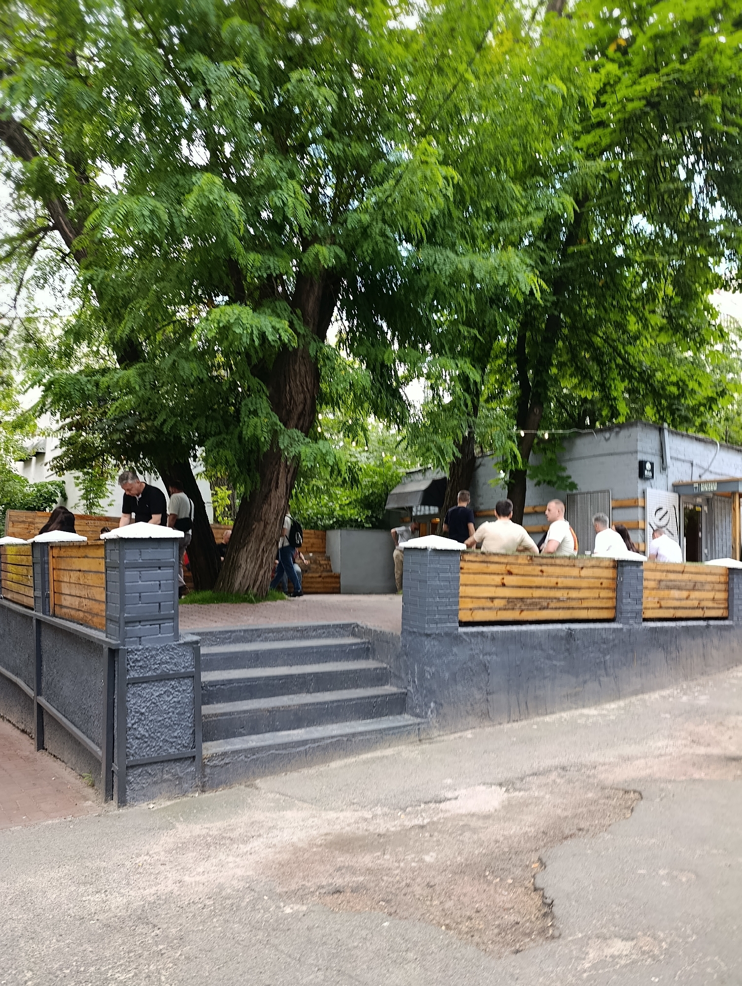 The image shows an outdoor seating area of a cafe or restaurant, likely in Kyiv, Ukraine, given the context. The area is set up with wooden benches and tables, separated by low wooden fences. Several people are seated at the tables, engaged in conversation or simply relaxing. The scene is framed by large trees with lush green foliage, suggesting a warm weather setting. In the background, a grey building with a dark awning and windows is visible, possibly the establishment itself. The ground is paved with asphalt. The overall impression is of a casual, urban outdoor dining experience.