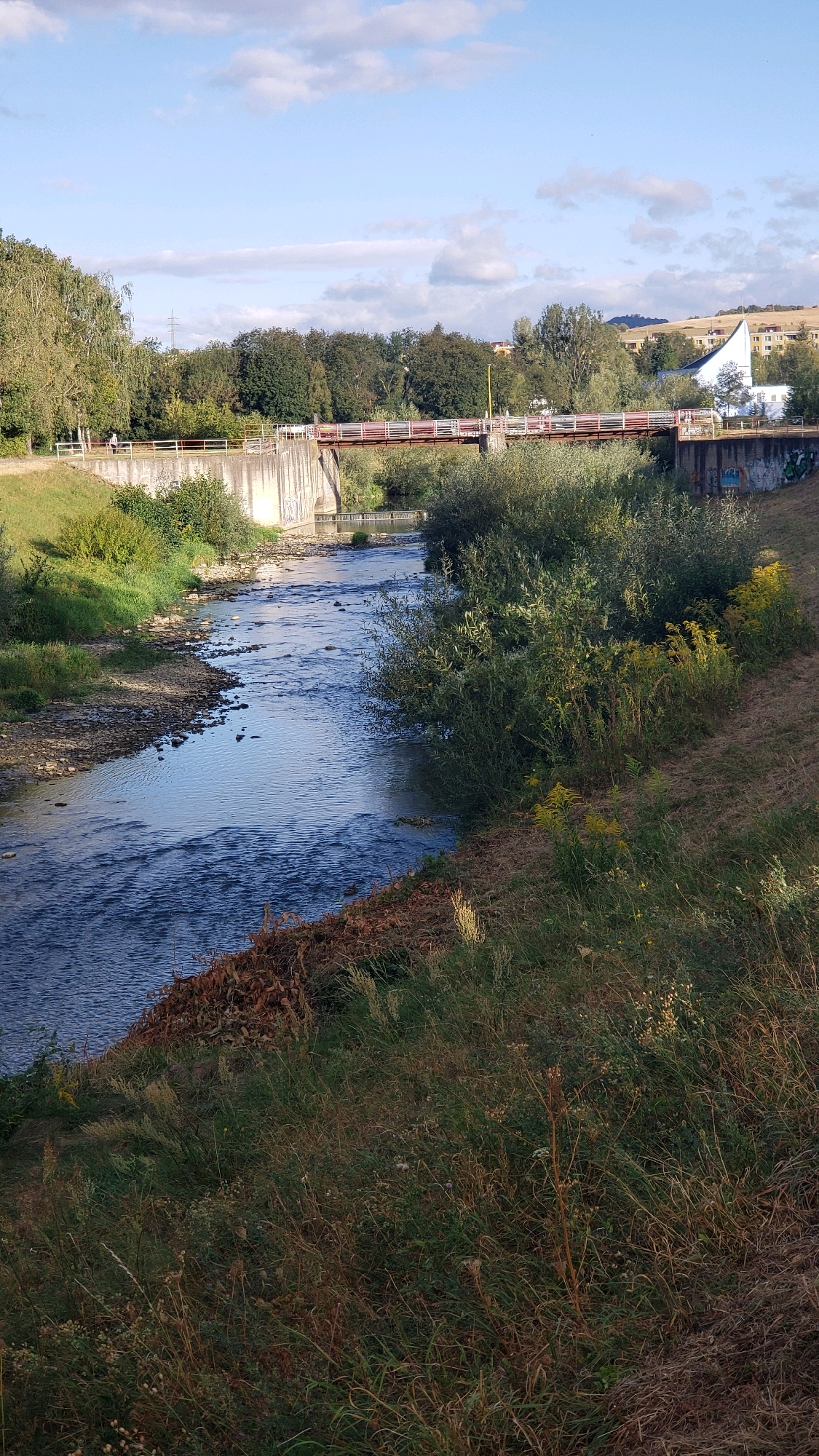 A narrow, concrete dam spans a river under a blue sky with scattered clouds. A red and white metal bridge crosses the river above the dam. The riverbanks are covered in green grass and trees. In the background, buildings and hills are visible. There is one person walking on the path next to the dam.
