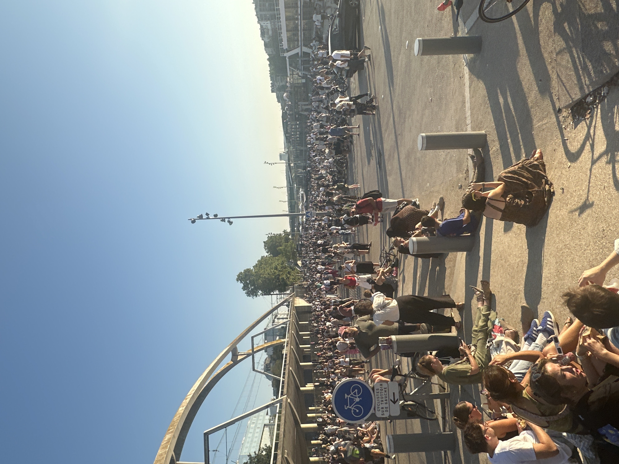 This is a wide-angle, eye-level, outdoor photograph of a large gathering of people on a paved area under a clear blue sky. The scene is likely Paris, France, given the architectural style of the overpass and the presence of what appears to be a boat moored on a body of water in the background.
In the foreground, several individuals are seated on the ground, some appear to be eating or relaxing. They are dressed in casual summer clothing. To the left, a blue sign with a white bicycle symbol and French text indicating "Cyclists on the sidewalk to the left" is visible. A bicycle is parked next to it.
Further back, a dense crowd of people is standing and walking, many wearing light-colored clothing. The crowd extends into the midground, where a dark-colored sedan is visible on the right side of the paved area. In the background, a multi-story structure, possibly a building or an elevated walkway, runs across the frame, supported by columns. A large arched bridge structure is also prominent on the left side of the image. Along the right side of the midground, a white boat or barge is moored. The setting sun casts long shadows across the paved surface. The overall atmosphere appears to be that of a casual public gathering on a warm day.