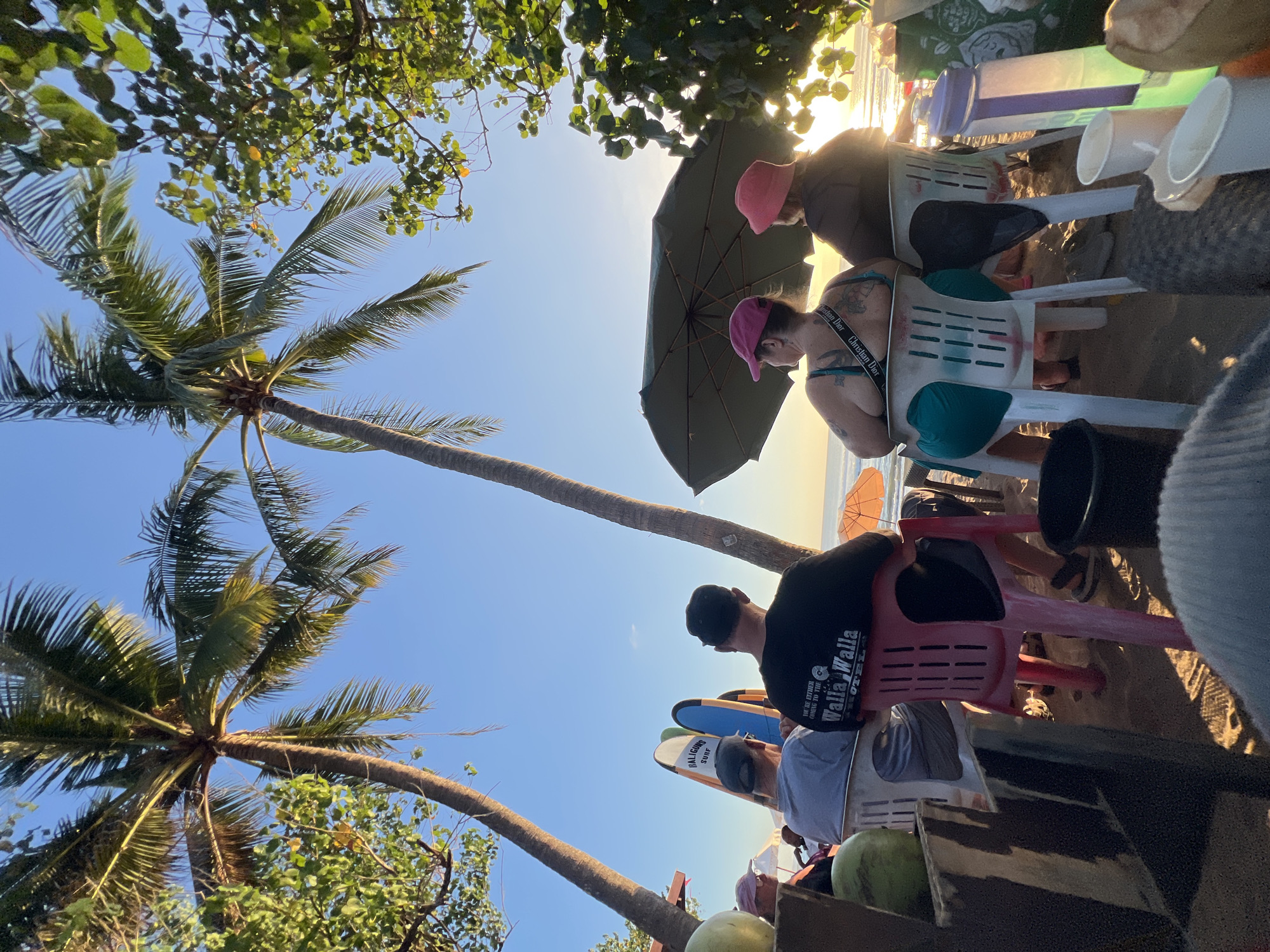 In Kuta, Indonesia, several individuals are seated on plastic chairs on a sandy beach, facing away from the camera. Palm trees frame the scene against a clear blue sky, with the setting sun casting a warm glow on the ocean. Two surfboards stand upright near the seated figures. A large green umbrella is open, providing shade. Among the people, some are wearing hats, and one individual has visible tattoos. Various objects, including coconuts, cups, a water bottle, and a bucket, are scattered around the seating area. The atmosphere appears relaxed, suggesting a casual gathering by the sea.