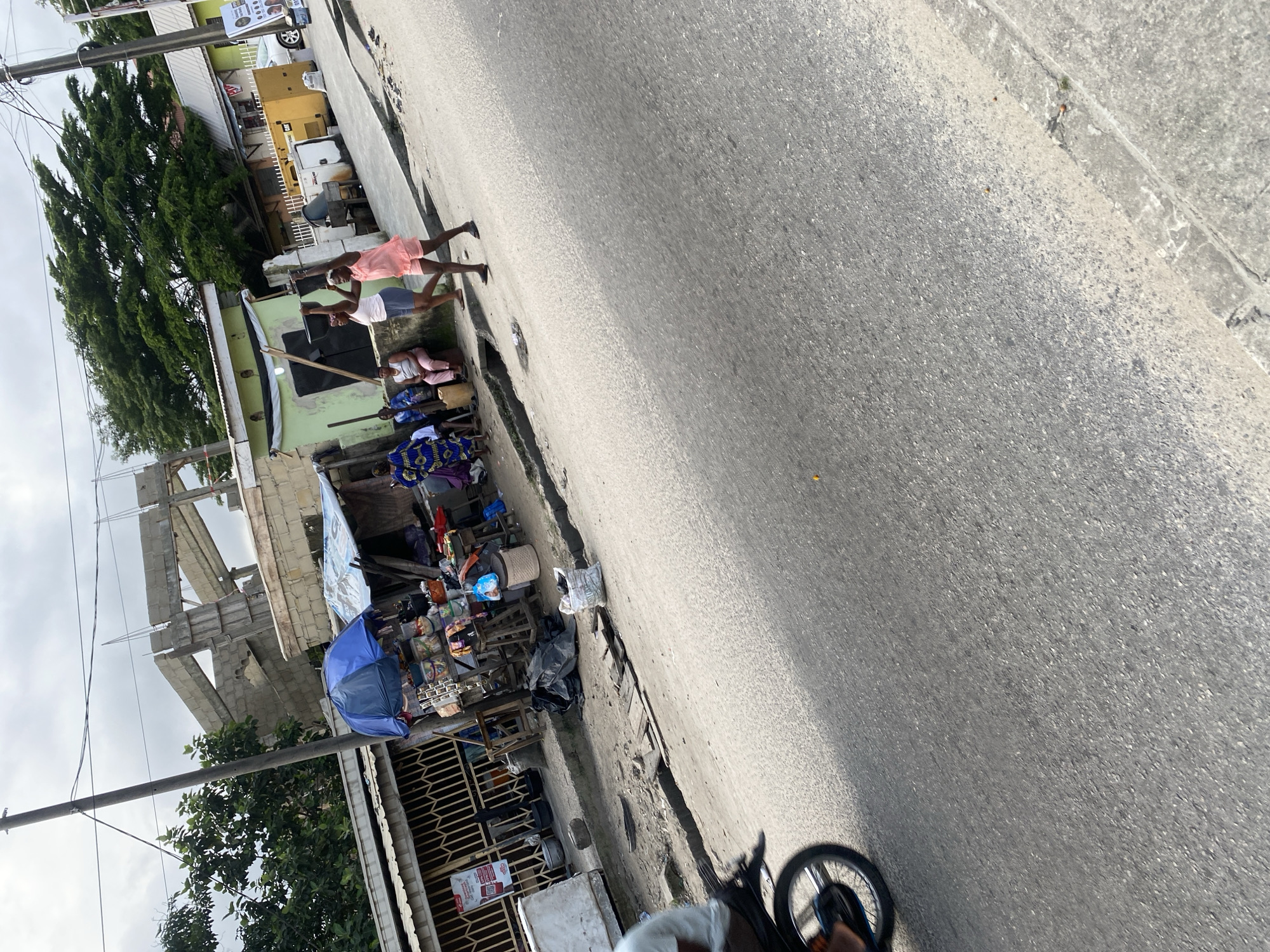 The image depicts a street scene with a market stall on the left and a road on the right. The market stall is set up under a covered area, with various goods displayed on shelves and tables. There are several people around the market stall, some of whom appear to be vendors and customers. In the background, there is a building under construction, with visible concrete structures and scaffolding. Several trees are also visible in the background, some of which are quite large and leafy. On the road, two young women are walking, one of whom is carrying a stick above her head. A motorcycle is visible in the foreground on the left, with its rear wheel turned towards the viewer. The overall scene suggests a busy urban environment, possibly in a developing country, with ongoing construction and a bustling market.