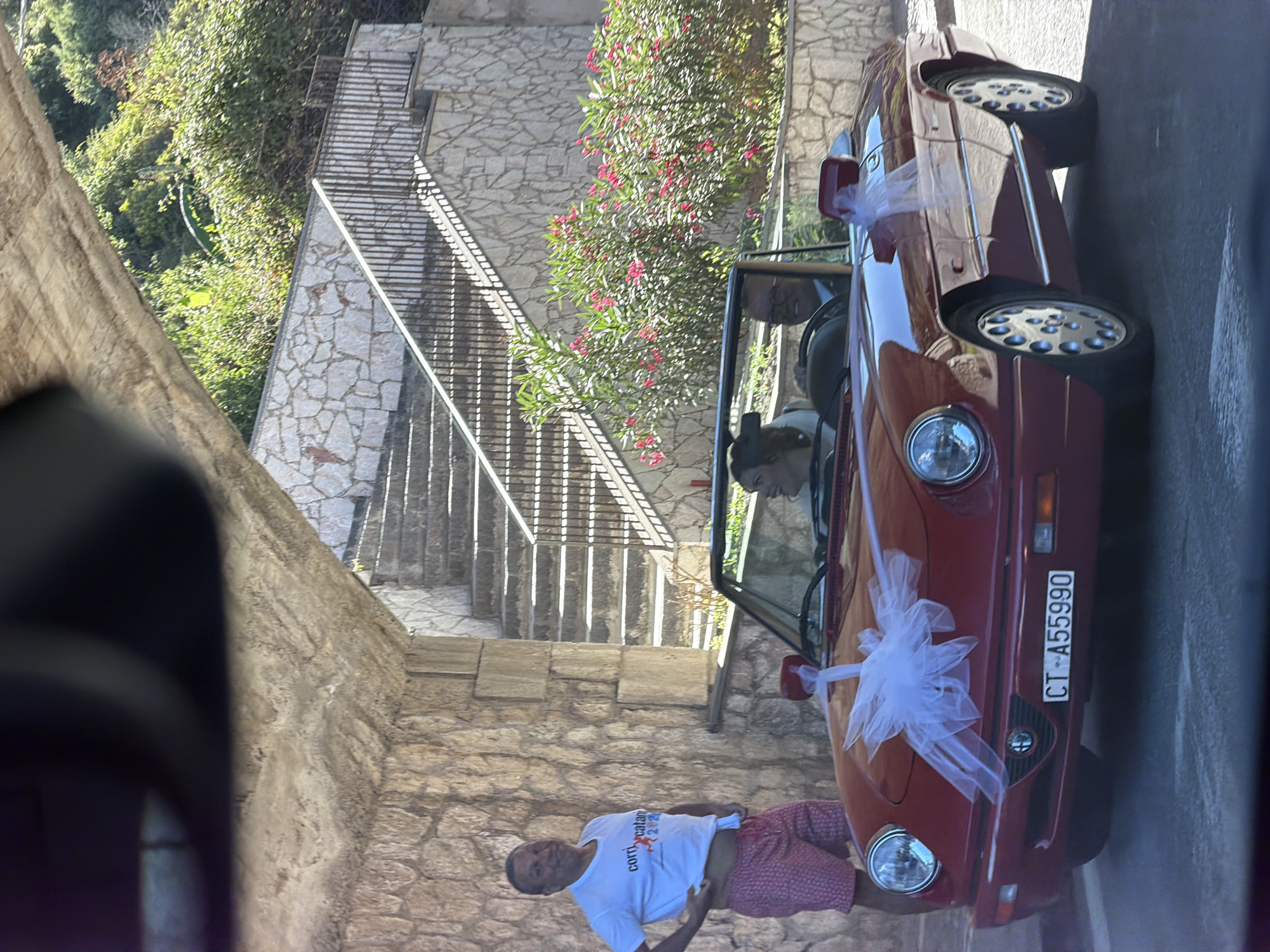 A man in a white t-shirt and red patterned shorts stands to the left of a red convertible, with stairs and stone walls in the background. Inside the convertible, a woman with dark hair and a white top is seated in the passenger seat, and a man wearing glasses and a suit is driving. The car is decorated with a white bow on the hood and a white ribbon on the passenger side mirror. The license plate reads "CT-A55990". Behind the car, there are blooming pink flowers and lush green foliage, with a stone-paved pathway leading up a set of stairs. The scene appears to be in a Mediterranean climate, possibly during a wedding celebration, and the architecture suggests a location in Taormina, Italy, known for its historical sites and scenic beauty.