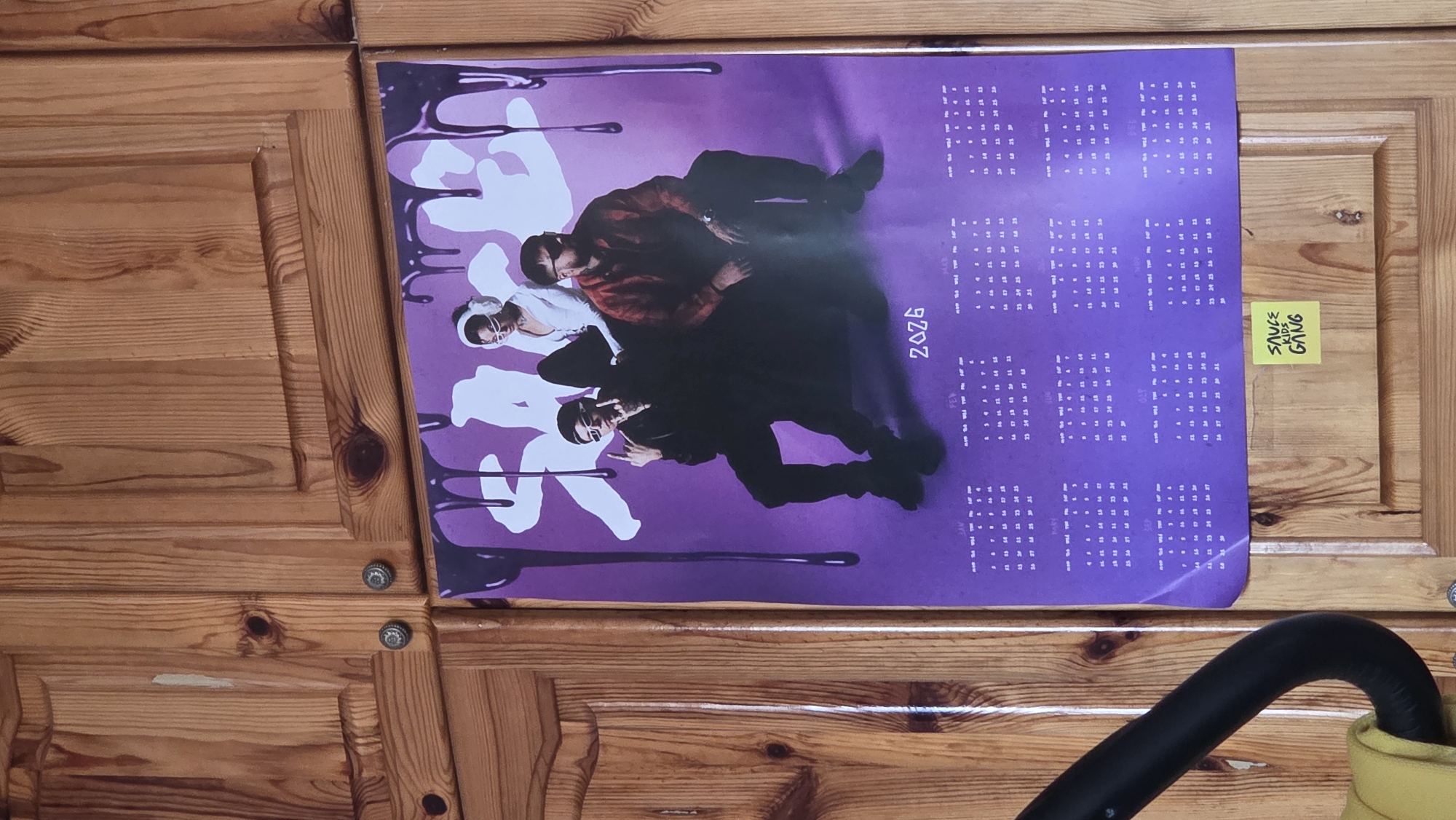 A full-shot image, rotated 90 degrees counter-clockwise, displays a vibrant purple poster of three young men hanging on a light brown wooden cabinet or wall. The wooden surface features distinct grain patterns and knots, with two small, metallic, decorative knob-like fixtures visible.
The poster itself is a calendar for the year 2026, with the numbers "2026" written vertically and reversed on the right side of the poster (due to the rotation). The background of the poster is a deep purple, adorned with stylized white and dark purple or black "drip" or splatter patterns.
The main subjects of the poster are three young men, possibly a music group or collective, posing in a cool, confident, and somewhat serious manner. They are dressed in casual, modern attire, including sunglasses, hoodies, and t-shirts. One man is giving a "rock on" hand gesture. Their expressions are unsmiling, contributing to an edgy aesthetic.
To the right of the poster, a small yellow sticky note is affixed to the wooden surface. The text on the sticky note, partially visible and oriented sideways, reads "SAUCE KIDS GANG". In the bottom right corner of the overall image, a curved, black object, possibly a handle or hose, extends into the frame. The scene suggests an indoor setting, likely a personal space given the casual display of the poster and sticky note.