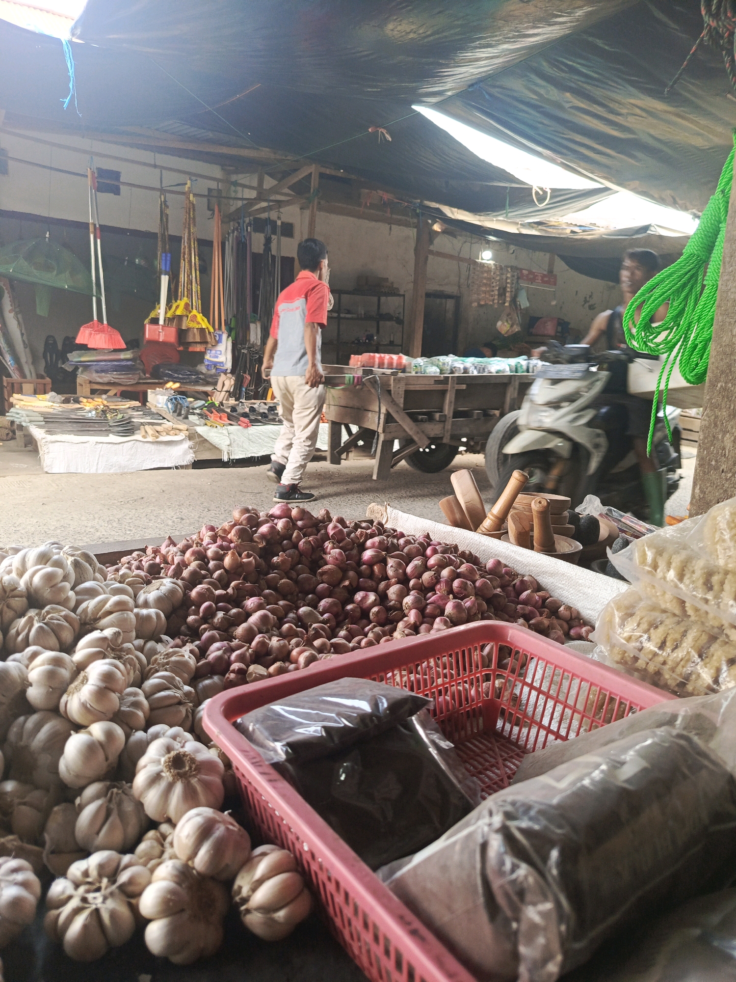 A medium shot captures an outdoor market stall in Megang Sakti I, Indonesia, under a tarp roof that lets in shafts of sunlight. In the foreground, a large pile of red onions sits next to a mound of garlic bulbs. To the right, a red plastic basket holds several plastic-wrapped packages of dark powder and a bag of light-colored snacks. Further back, various brooms and other cleaning tools hang from racks. A man in a red and gray shirt walks away from the camera towards the center of the frame, past a wooden cart laden with goods. In the background, another person is partially visible behind a motorcycle. The ground appears to be made of packed dirt or concrete.