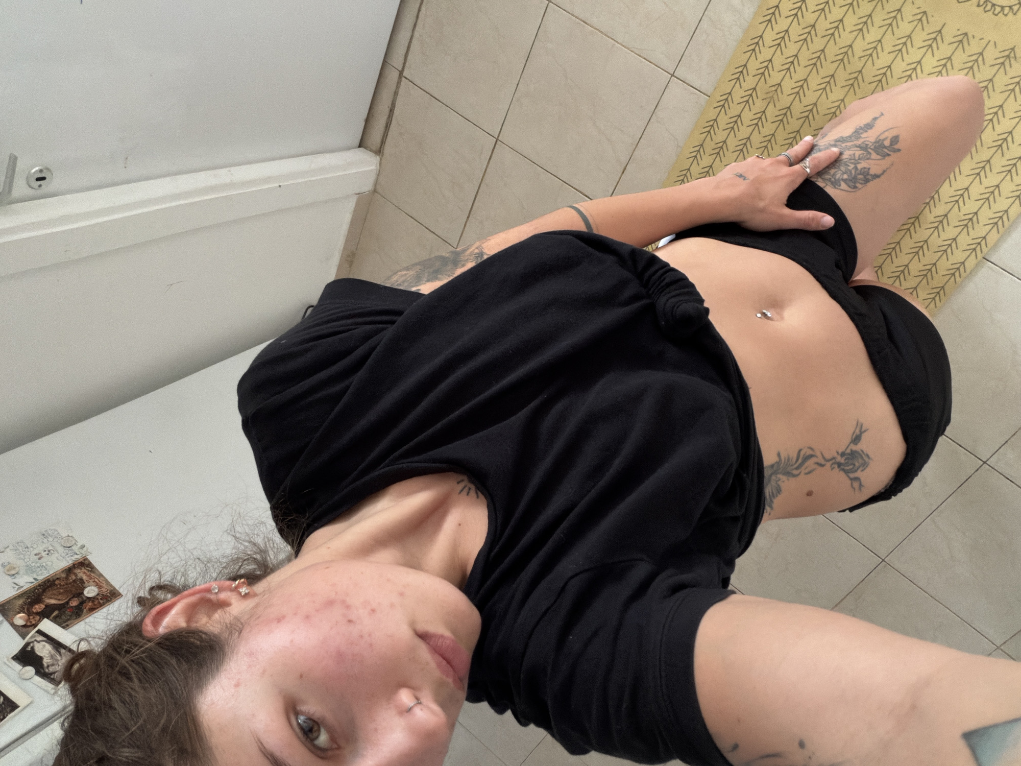 A high-angle, selfie-style photograph shows a person indoors. The person, likely female, is partially visible. They are wearing a black t-shirt and black shorts. Their abdomen is visible, and they have a pierced navel. Tattoos are present on their legs, torso, shoulder, and arm. The background consists of a tiled floor, a yoga mat with a geometric pattern, and a white cupboard/cabinet. The photograph appears to be taken in a domestic setting, possibly an apartment in Kiryat Bialik, Israel. Details in the image are insufficient to pinpoint an exact location within the city.