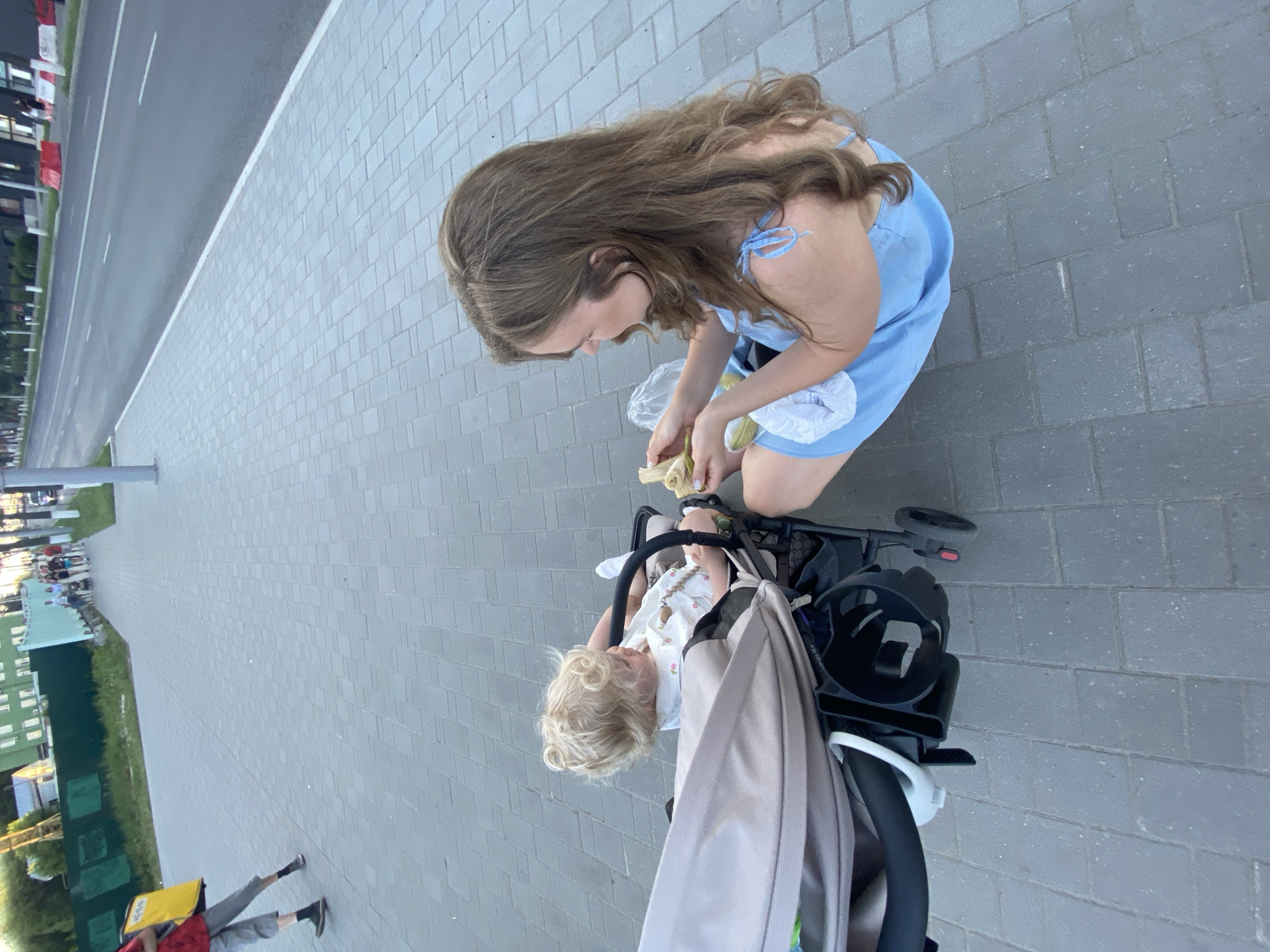 A woman in a blue dress is sitting on a sidewalk and feeding a baby in a stroller. The baby has blonde curly hair and is wearing a white outfit. The woman is holding pieces of a banana and offering them to the baby. The stroller is a double stroller, and a portion of the second seat is visible. The background shows a paved sidewalk, a street with cars, and some buildings. The scene appears to be outdoors in a public area. This image was taken on Tsvetova Street in the city of Vladimir, Russia.