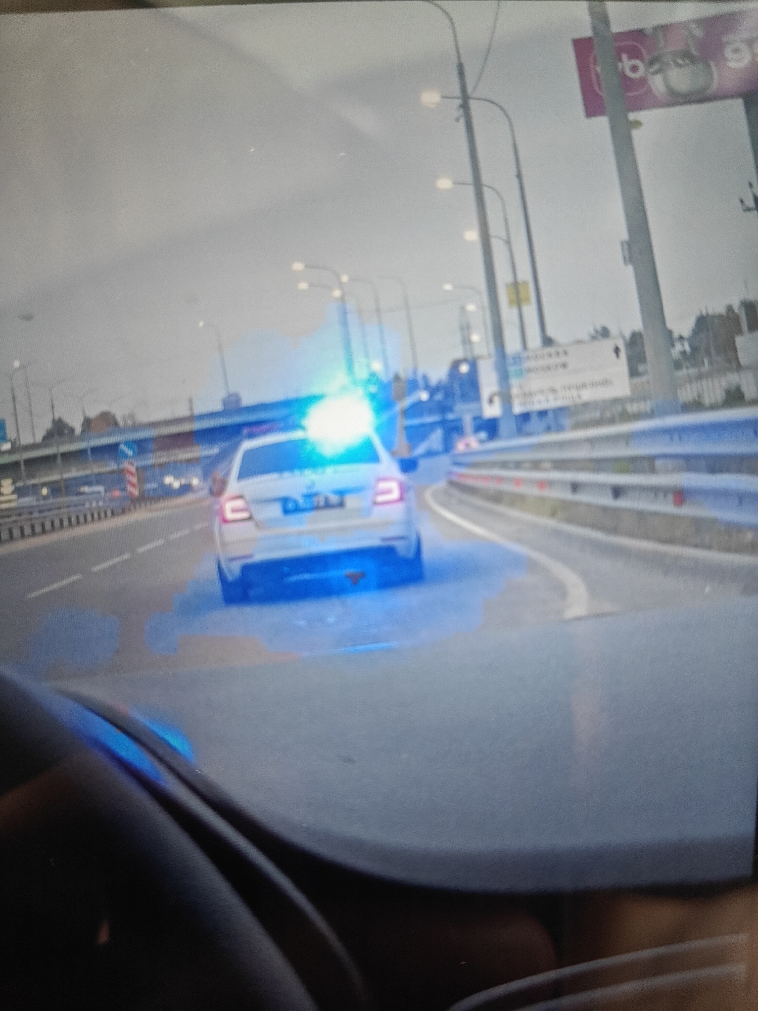 The image depicts a white police car with blue flashing lights driving on a highway. The car is in the middle lane of a multi-lane road, with a concrete barrier to the right and other vehicles and road infrastructure visible in the background. There is a sign on the right side of the highway with Cyrillic text and a logo that appears to be "b." The sky is overcast. The image appears to be taken from inside another vehicle, looking forward. Based on the road signs and general appearance, the location is likely a highway near Pushkino, Russia.