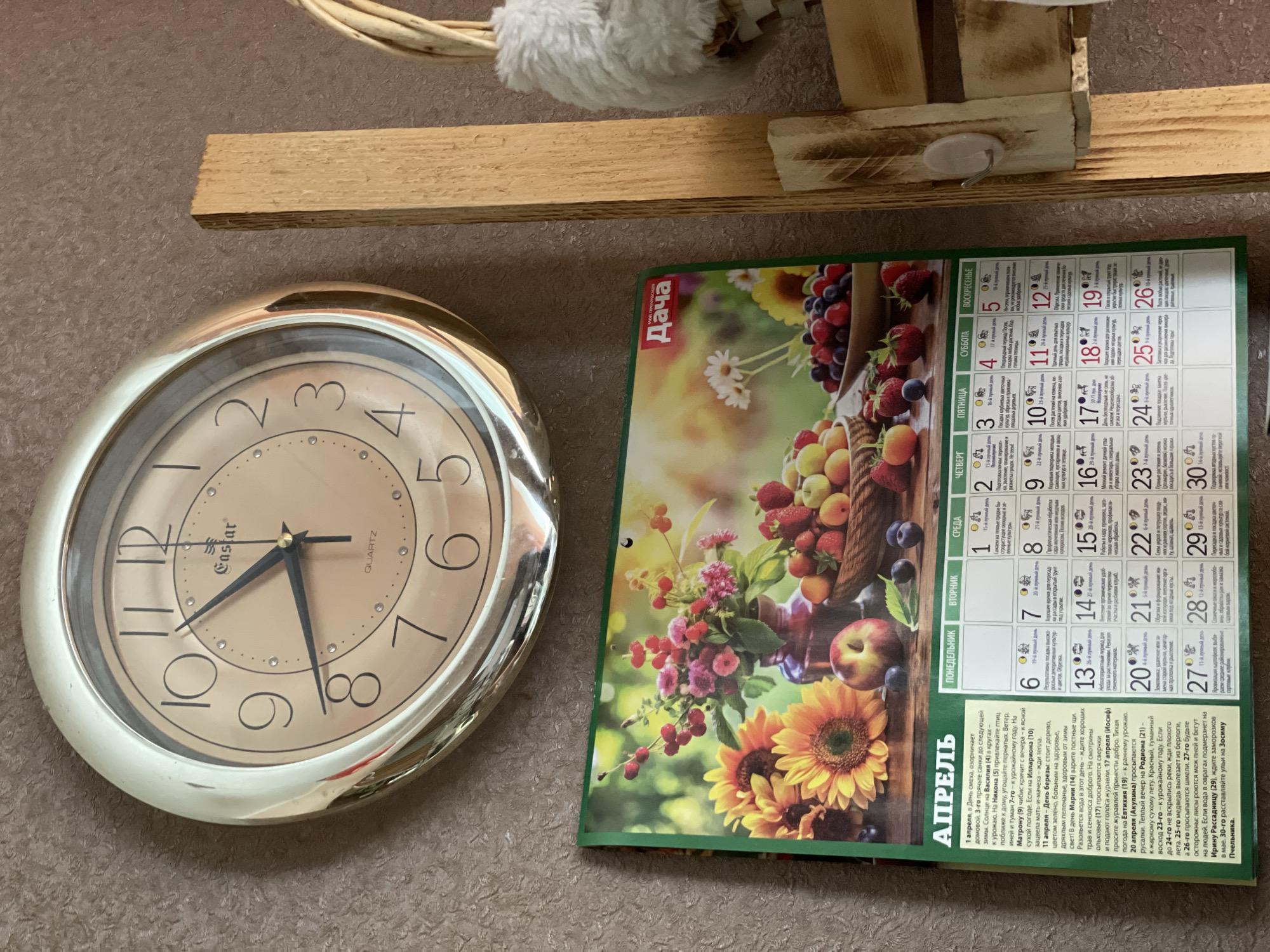 The image shows a wall with a clock and a calendar hanging on it. The clock has a silver frame and a beige face with black numerals. The hands of the clock indicate that the time is approximately 8:28. The calendar is open to the month of April and features an image of a basket of fruit and sunflowers. The calendar is written in Russian. There is also part of a wooden shelf with a decorative item above the clock and calendar. The wall is a neutral brown color. I cannot determine the location in the city of Zheleznogorsk, Russia, or any other geographic information, people, subjects, actions or interactions from the provided information.