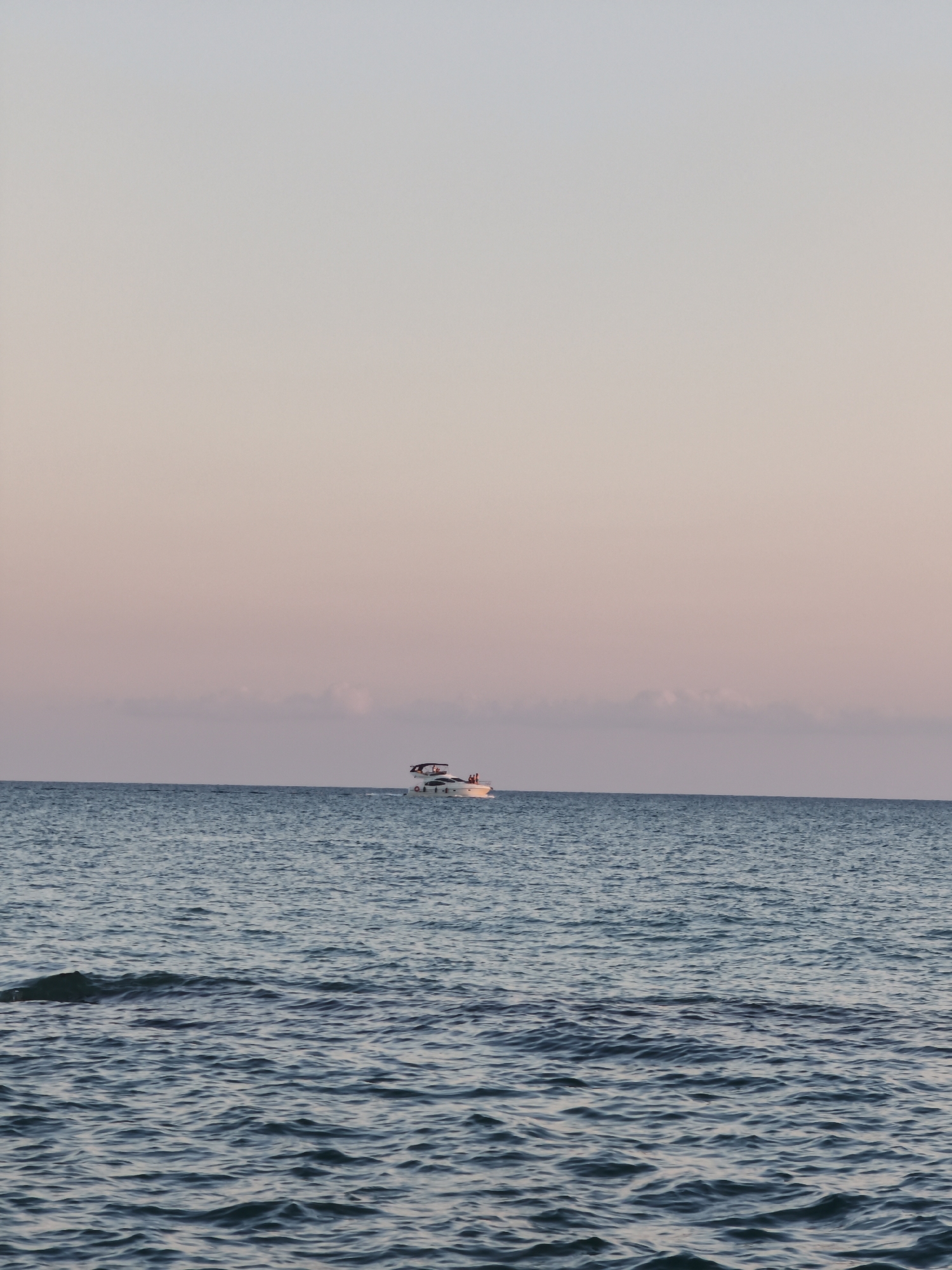 A wide shot captures a serene ocean scene under a pale pink and blue gradient sky, indicative of either sunrise or sunset. In the middle ground, a white yacht with several occupants is visible on the water. The foreground features the undulating surface of the sea, with gentle waves reflecting the soft light. The environment is that of an open body of water, likely the Black Sea off the coast of Sochi, Russia. The overall atmosphere is peaceful and tranquil.