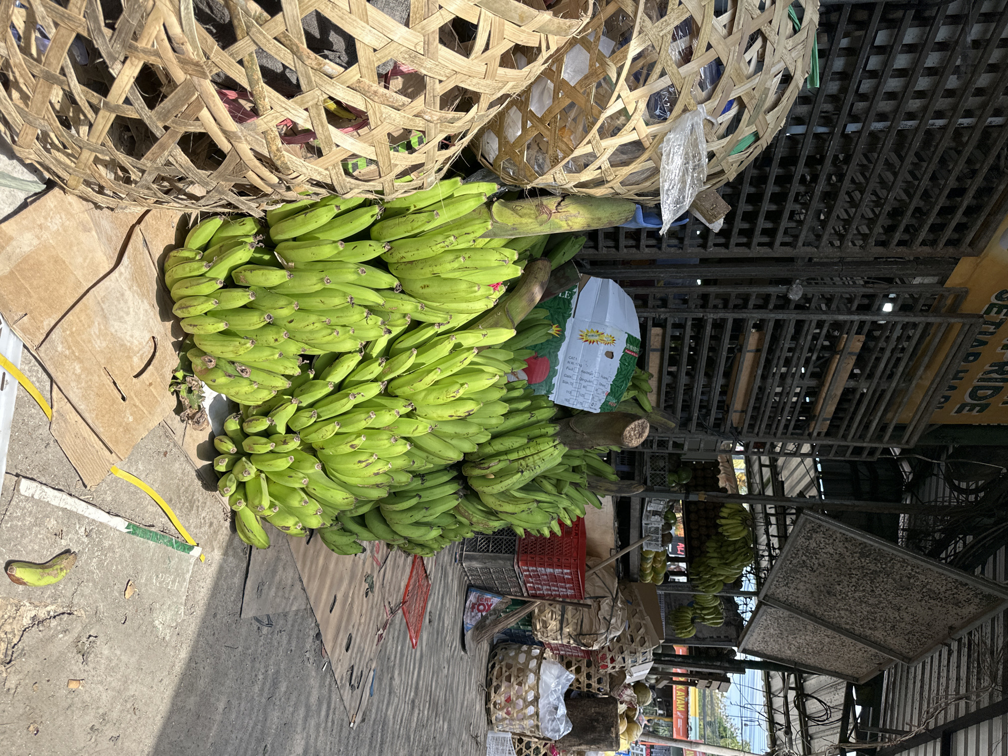This image depicts an outdoor market stall in Kuta Selatan, Indonesia. The stall displays a large quantity of green bananas, some of which are in woven baskets and others are piled on the ground. There are also coconuts and other fruits visible in the background. The market appears to be located on a street with a concrete surface and a covered structure above the stalls. A sign with text in Indonesian, including "AYAM," is visible on a building in the distance. The scene is brightly lit by sunlight, suggesting it is daytime. No people are visible in the frame.