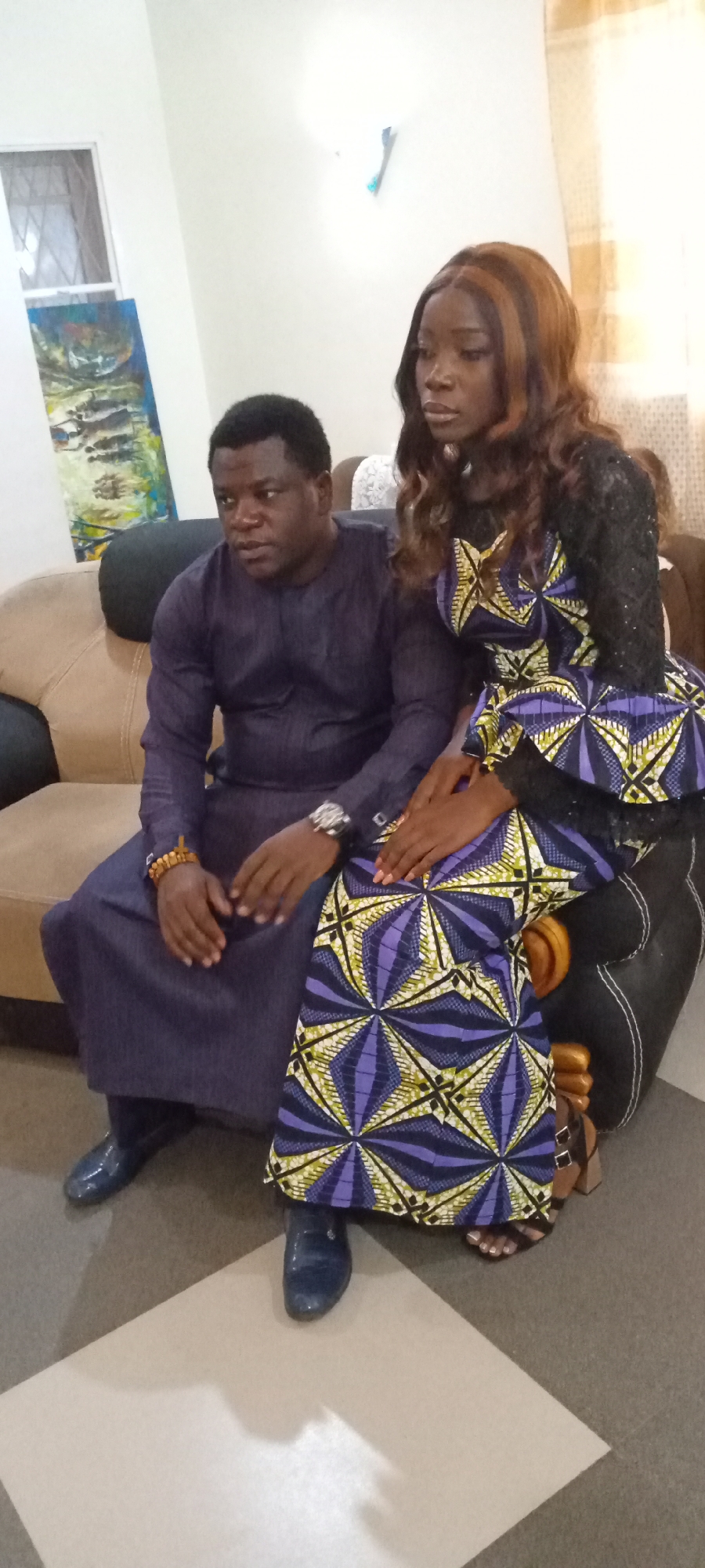 A medium shot shows a man and a woman indoors in Unknown, Unknown. The man, on the left, has dark hair and skin and wears a dark blue long-sleeved tunic and dark blue shoes. He is seated on a tan couch with black pillows. His hands are clasped. The woman, on the right, has long brown wavy hair and wears a black and purple patterned dress. Her hands are resting on her lap. They both appear to be looking forward. In the background, there is a painting on the wall and a white curtain. The floor is a beige tile.