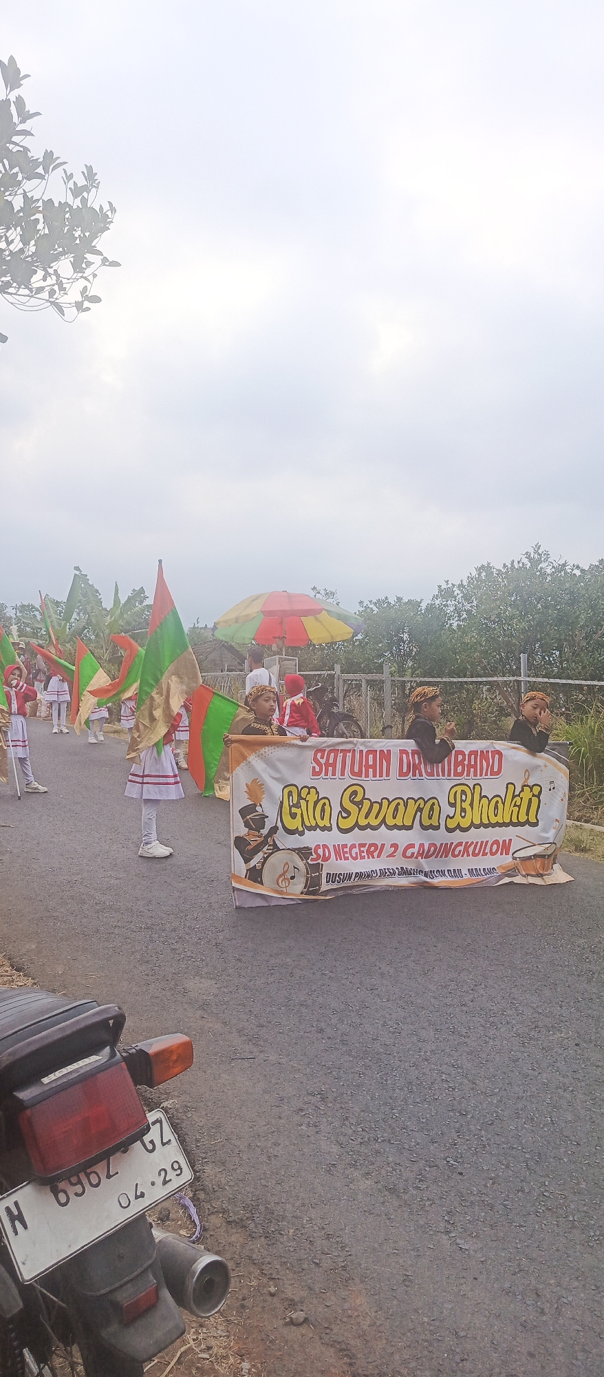 In Sumbersekar, Indonesia, a marching band parade is taking place on a paved road. The sky is overcast, and the scene is bordered by trees and vegetation on the right. In the foreground, the rear of a motorcycle with a license plate reading "N 6962 GZ 04.29" is visible. The parade participants, mostly children, are dressed in uniforms. Some are holding flags of red, green, and gold, while others are wearing traditional head coverings and dark outfits. A prominent banner across the road reads "SATUAN DRUMBAND Gita Swara Bhakti SD NEGERI 2 GADINGKULON" with an illustration of a drummer and musical notes. A colorful umbrella is positioned in the background. The overall atmosphere suggests a community event or celebration.