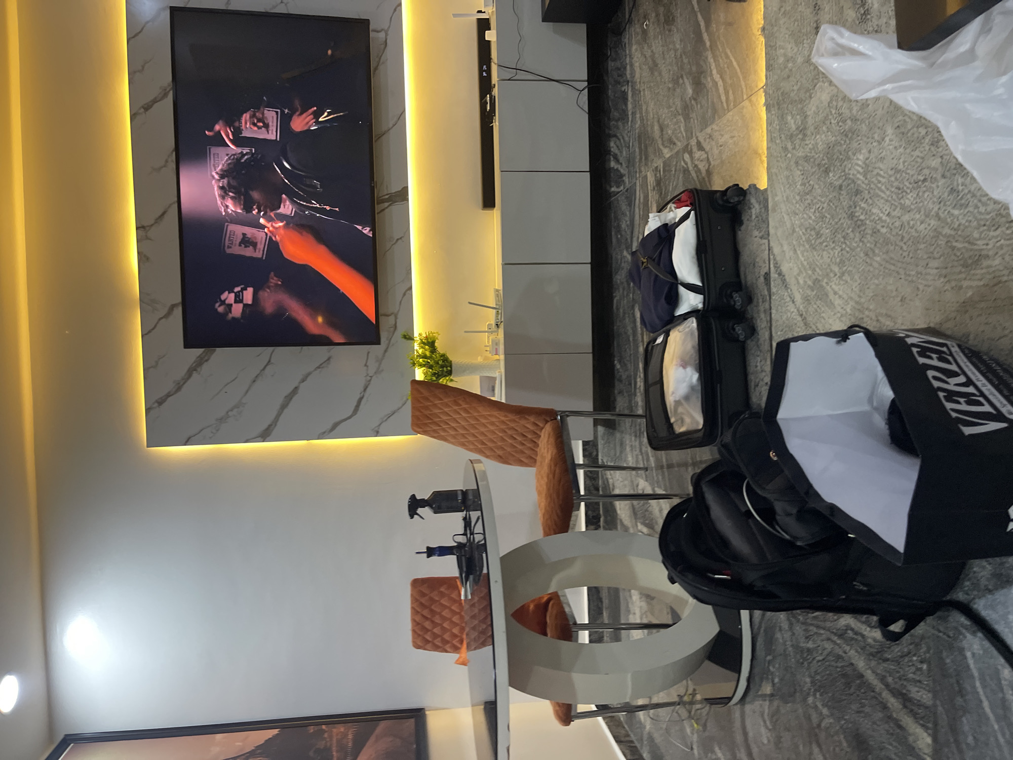 The image captures a modern interior space, likely a living and dining area in Tasha, Nigeria, featuring sleek design elements and warm lighting.
The central focus is a large flat-screen television mounted on a wall, displaying what appears to be a music video. The video shows several individuals, one prominent figure in the center with "WANTED" posters in the background, and another person with an orange-gloved hand near the bottom right. Below the television, a long, white media console or cabinet stretches across the wall, topped with a black soundbar and a small green plant in a white vase. The wall behind the TV and console features a white marble-effect panel with warm, hidden LED strip lighting running along its edges, illuminating the space.
To the left of the image (or lower in the original vertical orientation), a dining area is visible. It features a round, white table with a distinctive sculptural base. Two chairs with quilted, orange-brown upholstery are pulled up to the table. On the table, there's a black spray bottle and other small items.
On the floor, which has a grey, textured, or marble-patterned finish, several items are placed. A black rolling suitcase is open and packed with clothes, indicating recent travel or packing. Nearby, a black backpack and a larger black bag with white lettering, partially visible as "VERENDA," are also present. A crumpled white plastic bag lies further to the right. The room's overall aesthetic is contemporary, characterized by clean lines, accent lighting, and a blend of functional and decorative elements.