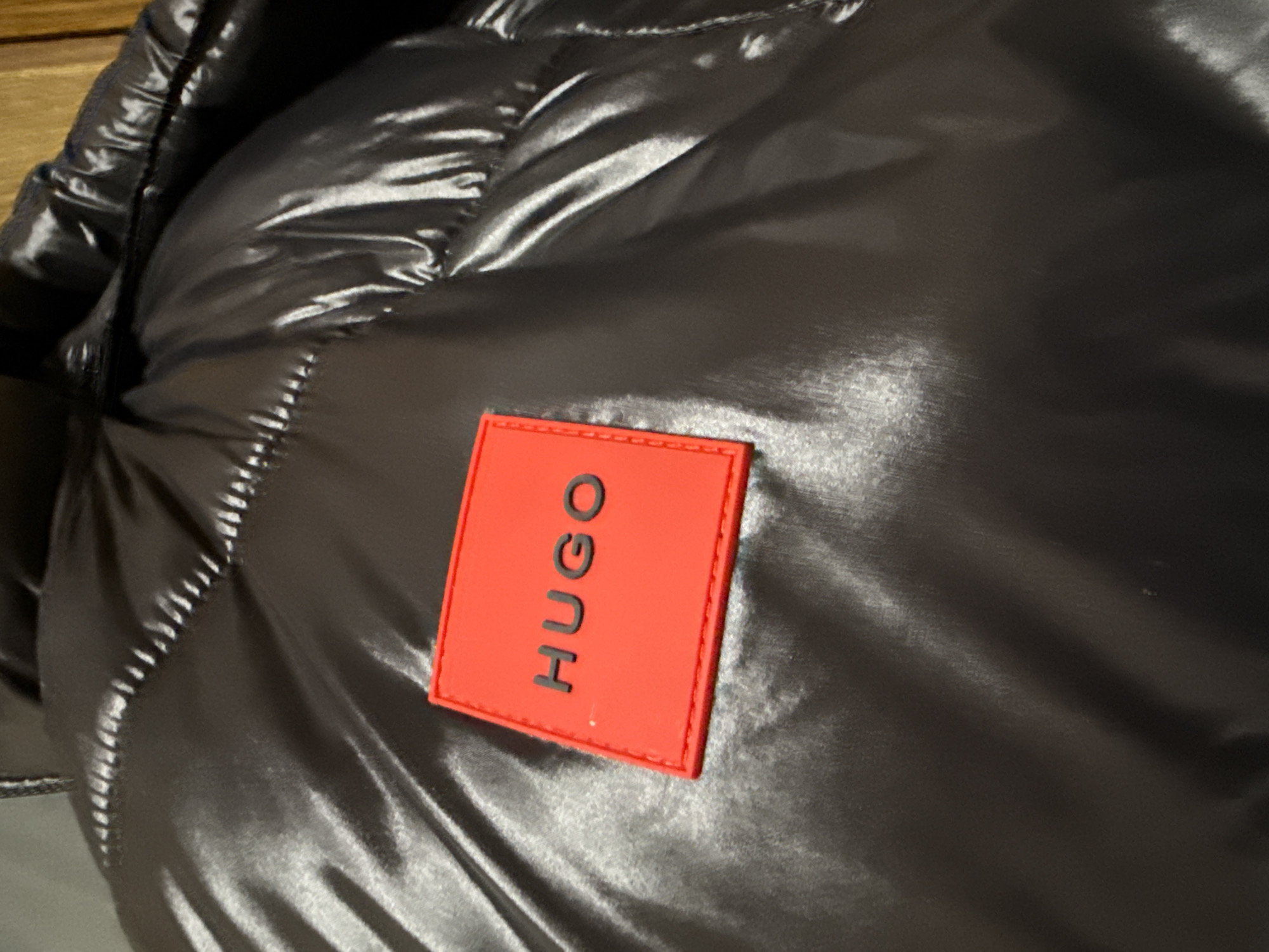The image shows a close-up of a shiny, dark-colored puffer jacket. A small, red rectangular patch with the word "HUGO" in black, vertically oriented letters is sewn onto the jacket. The fabric of the jacket has a quilted texture. The background is out of focus. The location in the city of Лабжанскі сельскі Савет, Belarus is not discernible from the provided image.
