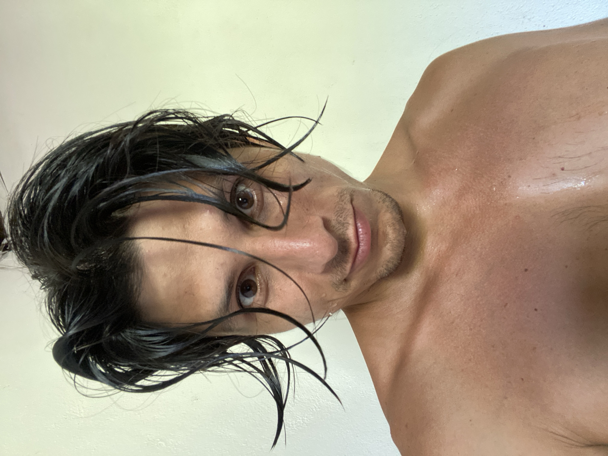 A close-up, chest-up, selfie-style photograph shows a person with dark, wet hair and brown eyes. The person's face is angled slightly towards the camera, with a faint smile. Their skin appears slightly flushed and is dotted with small beads of sweat, particularly on the chest and shoulders. The background is a plain, light-colored wall. The image is taken indoors, possibly in a residential setting in Los Velásquez, Venezuela.