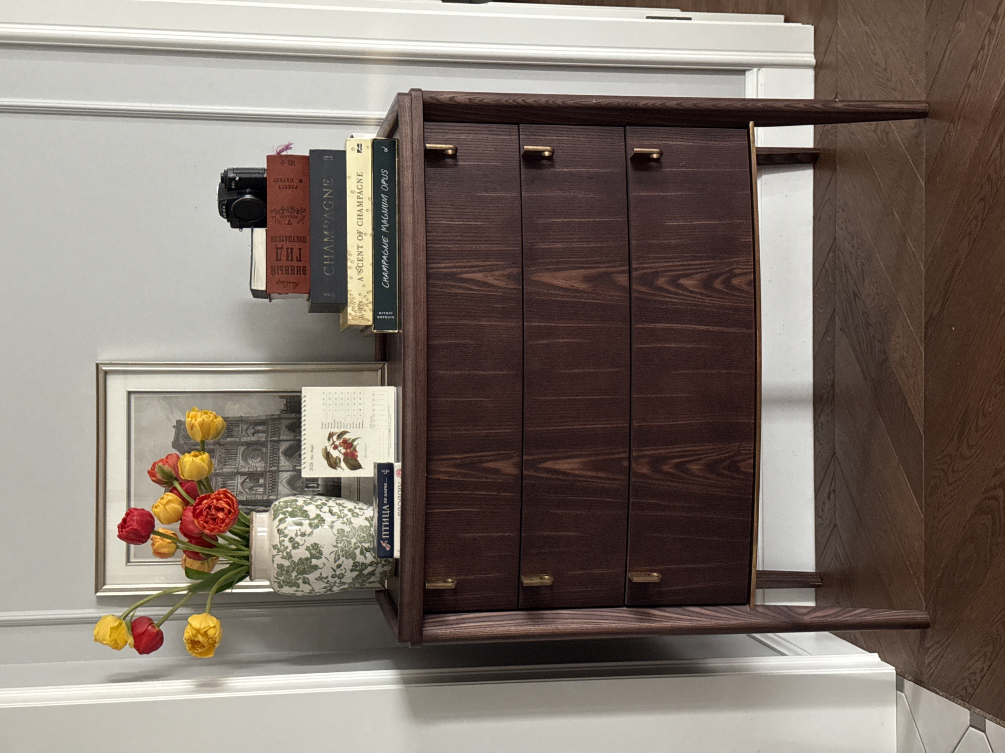 A low-angle shot presents a wooden sideboard positioned against a wall painted gray, which transitions to white near the top, adorned with decorative trim. The sideboard is divided into three vertical sections by its doors, each featuring a small, horizontal gold handle. Books and a black camera are stacked atop the sideboard. From left to right, the books include a camera, a brown book, "Champagne", "Ascent of Champagne", and "Champagne Magnum Opus". Adjacent to the sideboard, a silver-framed picture rests on the wall, accompanied by a vase filled with yellow and red tulips. The vase is decorated with a green leaf pattern. To the right of the sideboard, herringbone-patterned wooden flooring is visible. The books include "ПТИЦА по имени..." and "Гид ПОКУПАТЕЛЯ". There is no visible location within Moscow, Russia.