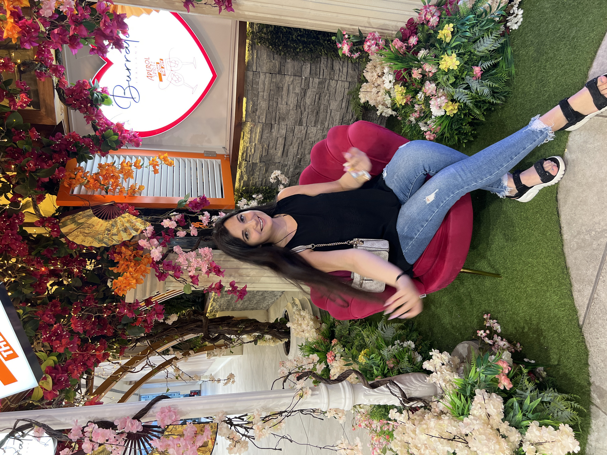 A young woman with long, dark hair sits in a plush pink chair amidst an elaborate floral display, a neon heart sign in the background. She wears a black sleeveless top and ripped blue jeans, accessorized with a small white purse and black sandals. The artificial greenery and flowers create a vibrant, immersive atmosphere, suggesting an indoor setting decorated for a festive occasion or restaurant ambiance. The scene appears to be an interior decorated with abundant artificial flora and a prominently displayed heart-shaped neon sign that reads "Burrata Italiana Aperol Spritz." The woman is positioned slightly off-center, looking towards the viewer with a smile. The floor is covered with artificial grass, and a light-colored pillar with floral embellishments is visible to her left. The overall impression is one of a carefully curated, cheerful environment.