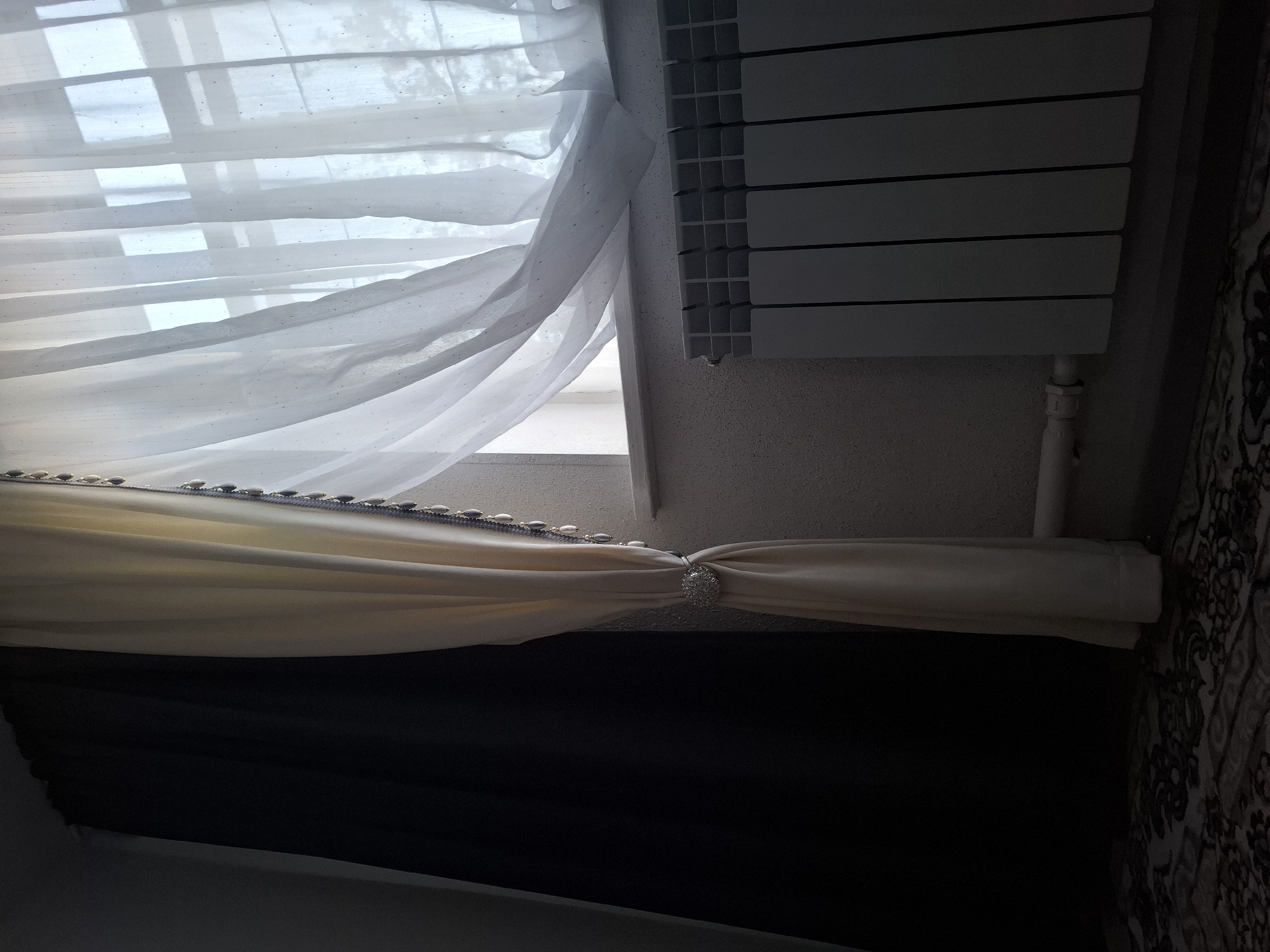This is an indoor photograph taken in Jeti-Ögüz, Kyrgyzstan, depicting a window area with curtains and a radiator. On the left side of the image, a dark, heavy curtain is drawn back, revealing a lighter, cream-colored curtain that is partially open. This cream curtain is held back by a decorative tie-back featuring a circular, sparkling brooch and a beaded trim along its edge. Beyond the cream curtain, a sheer white curtain with subtle, scattered embellishments is visible, obscuring the view outside the window but allowing light to filter through. The window frame appears to be white. Beneath the window, a white radiator with multiple vertical fins spans across the frame, connected by a pipe. The wall behind the radiator and window appears to be textured. The floor in the foreground is covered by a patterned rug, with the pattern appearing to be in shades of dark brown and beige. The lighting in the room is dim, with light primarily coming from the window. There are no people or animals present in the image.