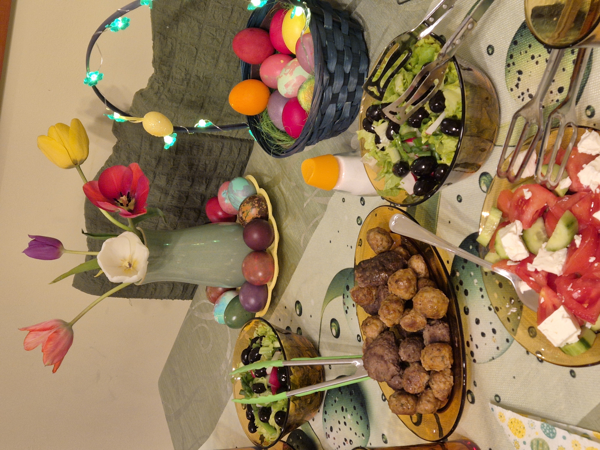 The image shows a table spread with a variety of food items and Easter decorations. There are two bowls of salad, one with olives and lettuce, and another with tomatoes, cucumbers, and feta cheese. There is a plate of meatballs. A vase with tulips and a basket full of colored eggs are also present. The location is unknown.