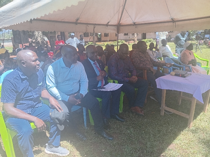 An outdoor gathering is depicted in Idakho South ward, Kenya, under a white tent canopy. Approximately 20-30 individuals, primarily adults, are visible on a grassy area. In the foreground, five men are seated on bright green plastic chairs, facing right. From left to right: a bald man in a dark blue polka-dotted shirt and jeans, holding a dark cap; a bald man in a light blue collared shirt, holding a dark object; a man in a dark suit with a red tie, holding documents; a man in a dark patterned long-sleeved shirt; and another man in a dark patterned long-sleeved shirt. To the right of these individuals, a wooden table covered with a light purple tablecloth holds a black microphone, a dark smartphone, and a small black object. A woman in a light top and patterned skirt is partially visible, seated on a light blue plastic chair further to the right. In the background, more attendees are visible, both seated and standing, with trees and a light-colored building discernible behind them. The individuals in the foreground appear attentive, suggesting observation of an ongoing event or speaker located off-frame to the right.