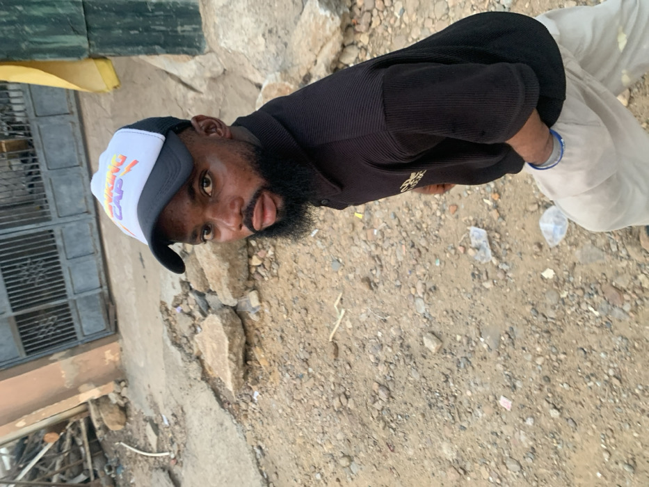 A man wearing a black ribbed shirt, white pants, and a white and black baseball cap with "Viking Cap" printed on it, stands outside on rocky terrain. His beard is dark and full. He is looking directly at the camera with his tongue slightly protruding. In the background, there's a building with a fenced window area, a patch of yellow material, and scattered debris on the ground.