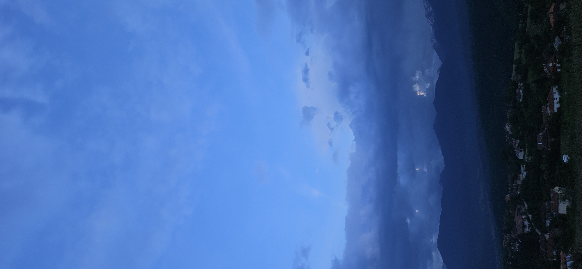The image displays a serene landscape captured during twilight or early evening. The sky, a dominant feature, presents a deep, rich blue hue, fading into darker, more overcast shades towards the right side of the frame. Soft, wispy clouds are scattered across the brighter blue expanse, while denser, darker cloud formations hug the distant mountain range.
A prominent, dark mountain range stretches horizontally across the mid-ground, silhouetted against the dusky sky. Its rugged peaks are partially obscured by the receding daylight and the gathering clouds. Towards the lower right portion of the image, a small village is nestled at the foot of these mountains. Scattered houses with reddish-brown tiled roofs are visible, interspersed with dark trees and patches of green land, all enveloped in the dim, evening light. A few faint, localized glows or reflections can be observed within the clouds near the mountains, hinting at a hidden light source or atmospheric effects.
The overall scene conveys a sense of peaceful tranquility as day transitions into night in a mountainous, rural environment. No specific activity or people are visible. There is no discernible text in the image. The setting is Gutsal, Bulgaria.