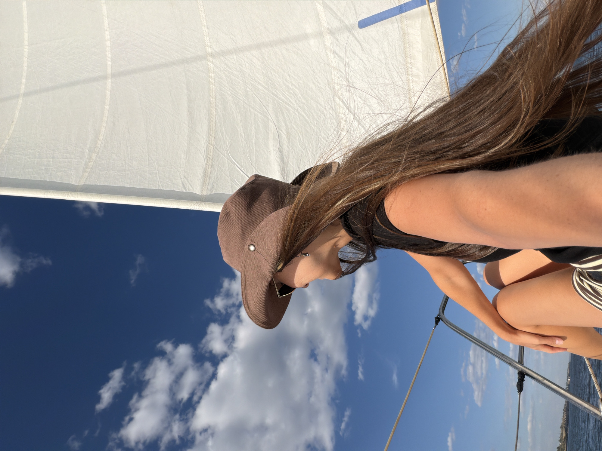 A young woman with long, brown hair, wearing a brown hat and sunglasses, is seen from the side as she sits on a sailboat. Her hair is blowing in the wind. She is wearing a black top and patterned shorts. The sailboat's white sail is visible, as is the blue sky with scattered clouds. The water and a distant shoreline are also visible. This image was captured near the city of Kamyshin, Russia.