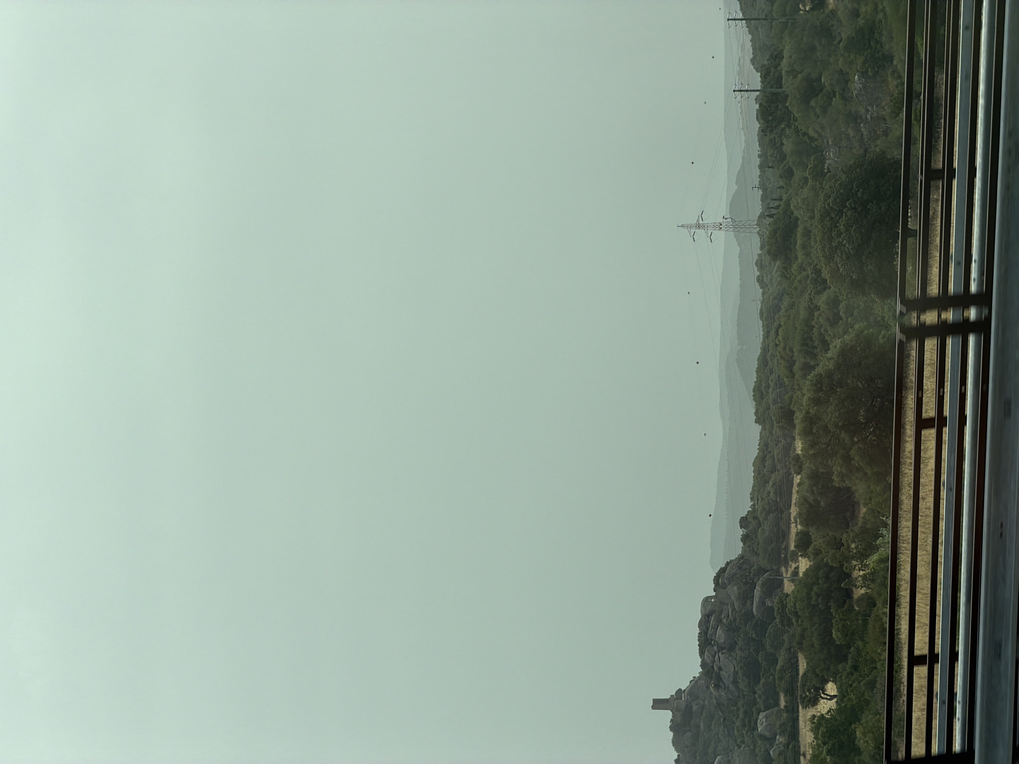 The image depicts a scenic view of the Sardinian landscape near Olbia, Italy. In the distance, a medieval tower stands atop a rocky outcrop, a testament to the region's history. The terrain is characterized by rolling hills covered in dense green vegetation, typical of the Mediterranean maquis. A prominent power line pylon rises above the trees, indicating the presence of modern infrastructure amidst the natural beauty. In the foreground, a railing of a bridge or overpass is visible, suggesting the photograph was taken from a moving vehicle, likely a car on a road. The sky is overcast, casting a soft, diffused light over the entire scene. The overall impression is one of a vast, sparsely populated rural area with a mix of historical and contemporary elements.