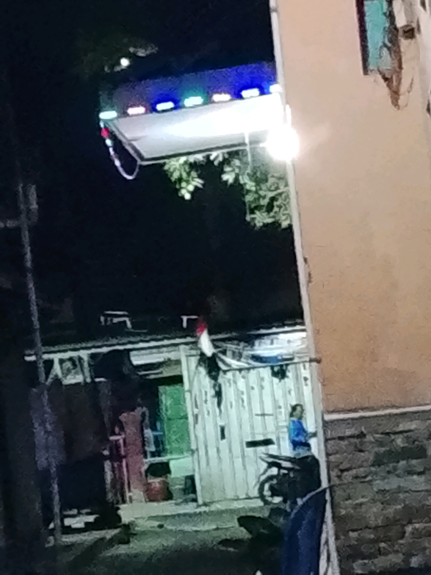 The image depicts an outdoor scene at night in Ujungrusi, Indonesia, with a focus on a makeshift stall or cart illuminated by a row of colorful LED lights. The stall, possibly a food or drink vendor, is covered and supported by poles, with some greenery hanging from it. In the background, a building with a textured, light-colored wall is visible. To the left of the stall, a dark, narrow alley or passage can be seen. On the right side of the image, a person wearing a blue shirt is standing next to a motorcycle. The ground appears to be paved with bricks or cobblestones. The overall impression is of a modest, potentially informal commercial setup in a residential or mixed-use urban area.